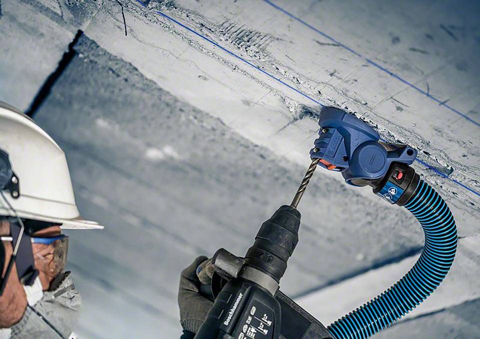 Un ouvrier portant un casque et des lunettes de protection fore un plafond en béton avec une perceuse bleue. Un dispositif d'aspiration élimine la poussière.