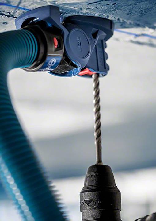 Un perceuse travaille sur un plafond, avec un tuyau d'aspirateur bleu branché pour capturer la poussière.