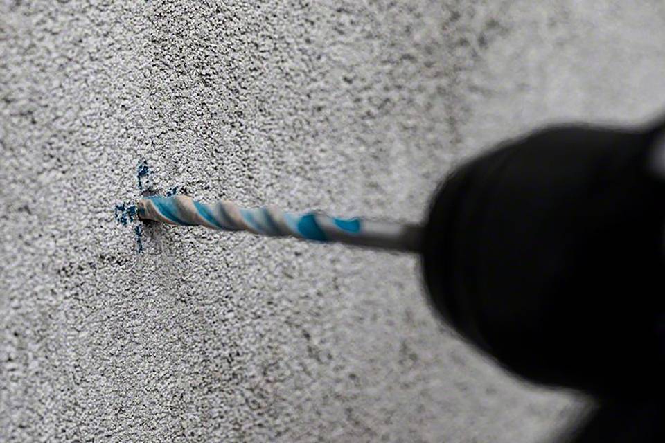 Un foret s'enfonce dans un mur de béton gris, de la poussière jaillit du trou de forage.