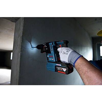 Une personne utilise un marteau perforateur à batterie Bosch bleu sur un mur en béton à l'intérieur. Elle porte un gant de sécurité.