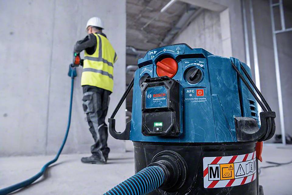 Un ouvrier en tenue de protection utilise un aspirateur industriel sur un chantier de construction. L'aspirateur est au premier plan.