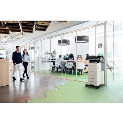 Environnement de bureau avec des murs en verre, où des personnes sont réunies en réunion. Deux personnes passent devant un imprimante multifonction.