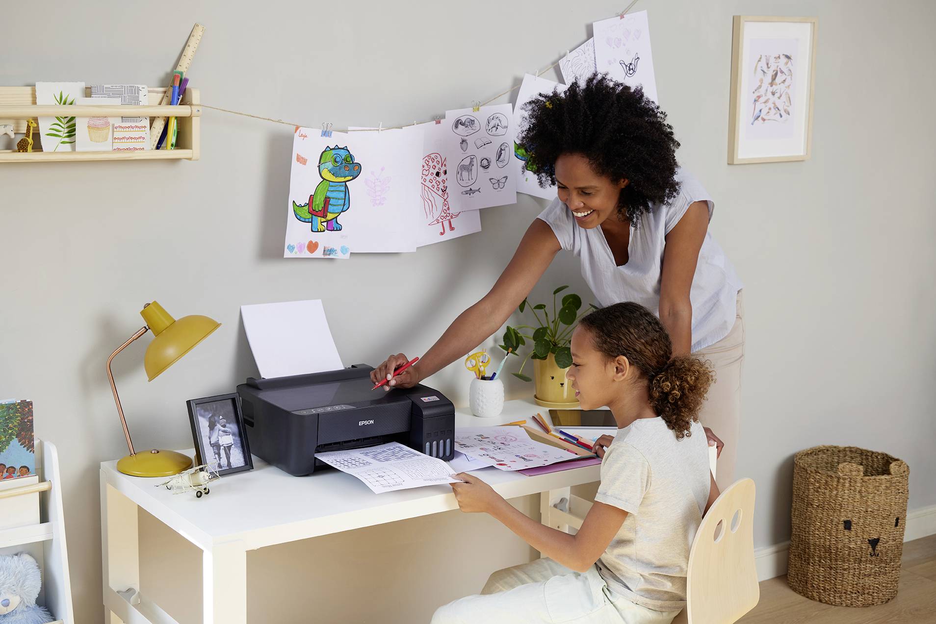Une femme aide un enfant à travailler sur un bureau. Des dessins sont accrochés en arrière-plan, et une imprimante est posée sur la table.