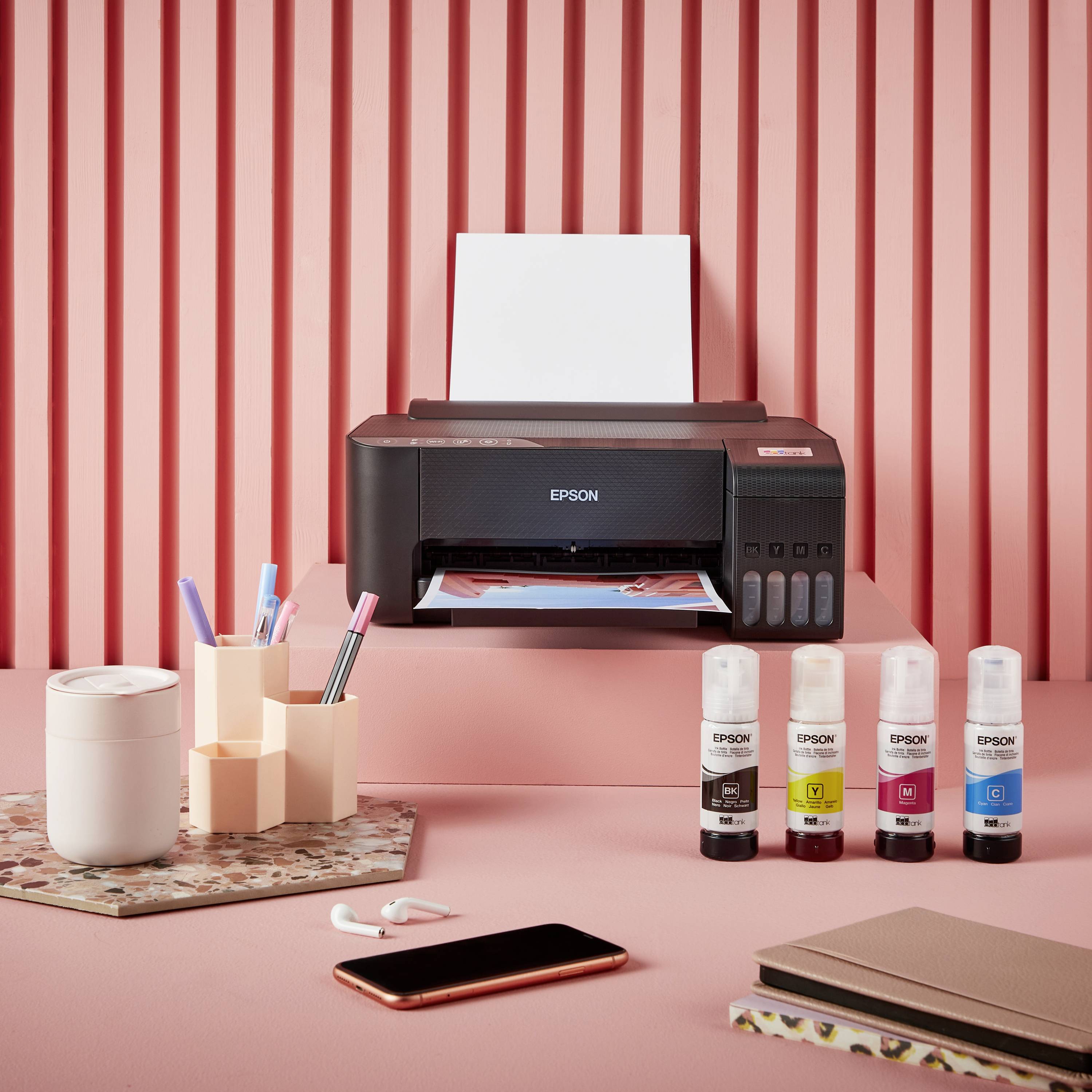 Une imprimante sur un bureau avec des bouteilles d'encre à côté. En arrière-plan, un mur rose avec des rayures verticales.