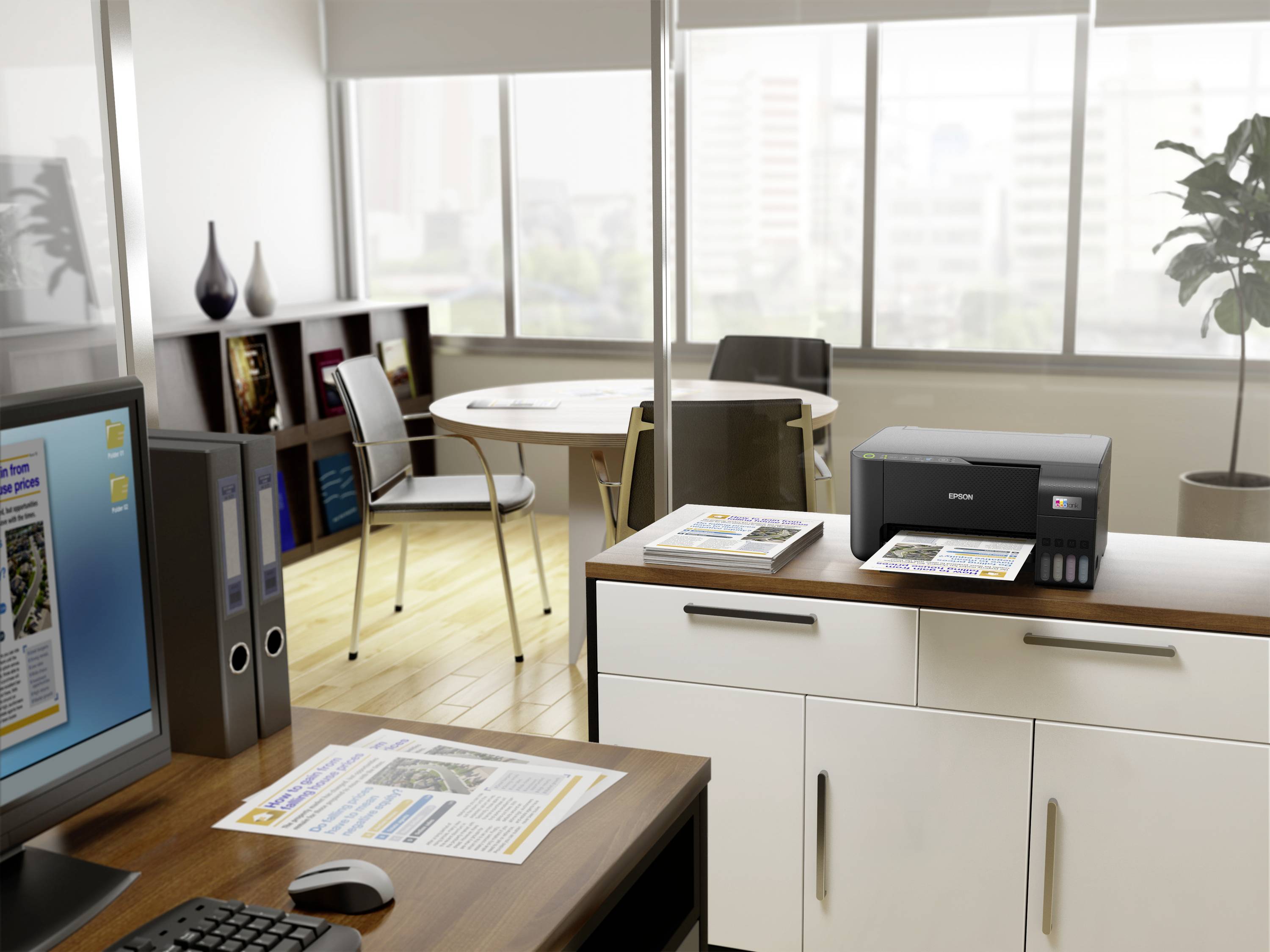 Un bureau avec un bureau, un ordinateur et une imprimante. Des impressions colorées sont posées sur l'écran et la table. Au fond, une table ronde et des étagères.