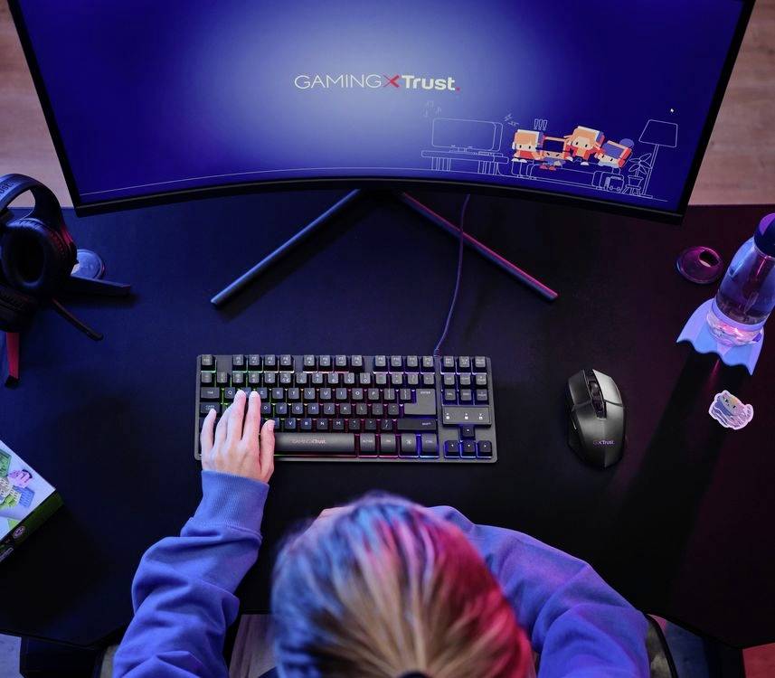 Personne assise à un poste de travail informatique avec un écran de gaming incurvé. Sur l'écran, 'GAMING XTrust'. Accessoires tels que souris et casque audio visibles.