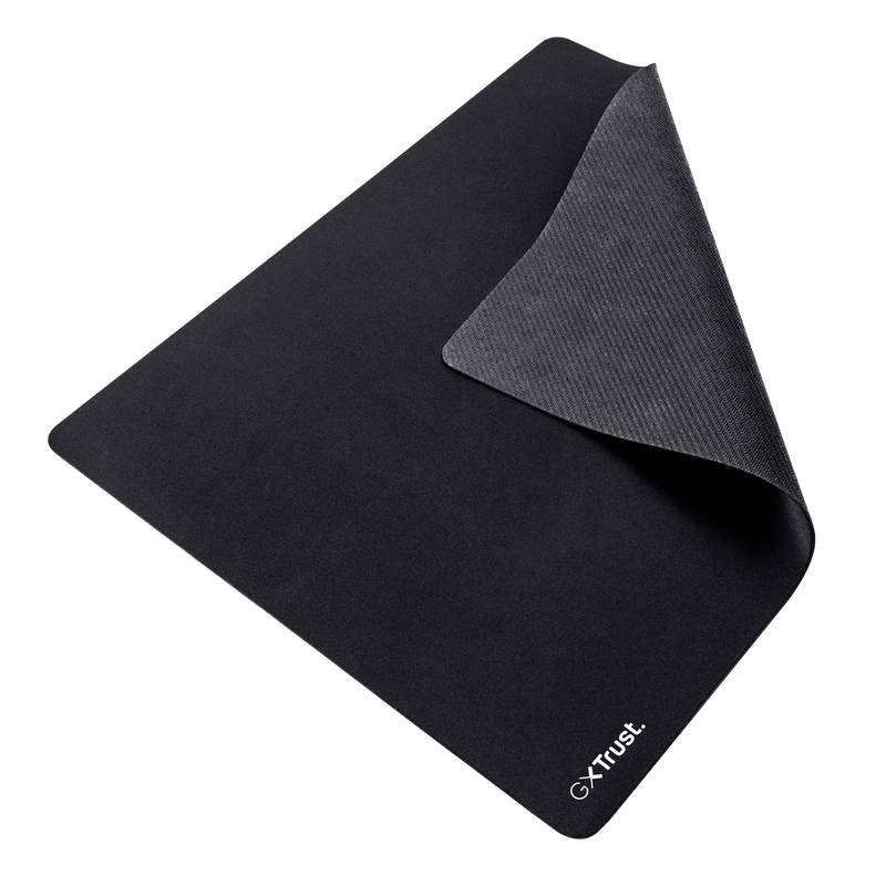 Tapis de souris noir avec un coin légèrement surélevé, montrant la couche grise inférieure. Logo 'Trust' en blanc en bas à droite.