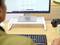 Une personne est assise devant un bureau, regardant un écran avec du texte. Sur la table se trouvent un clavier, une caméra et une tasse.
