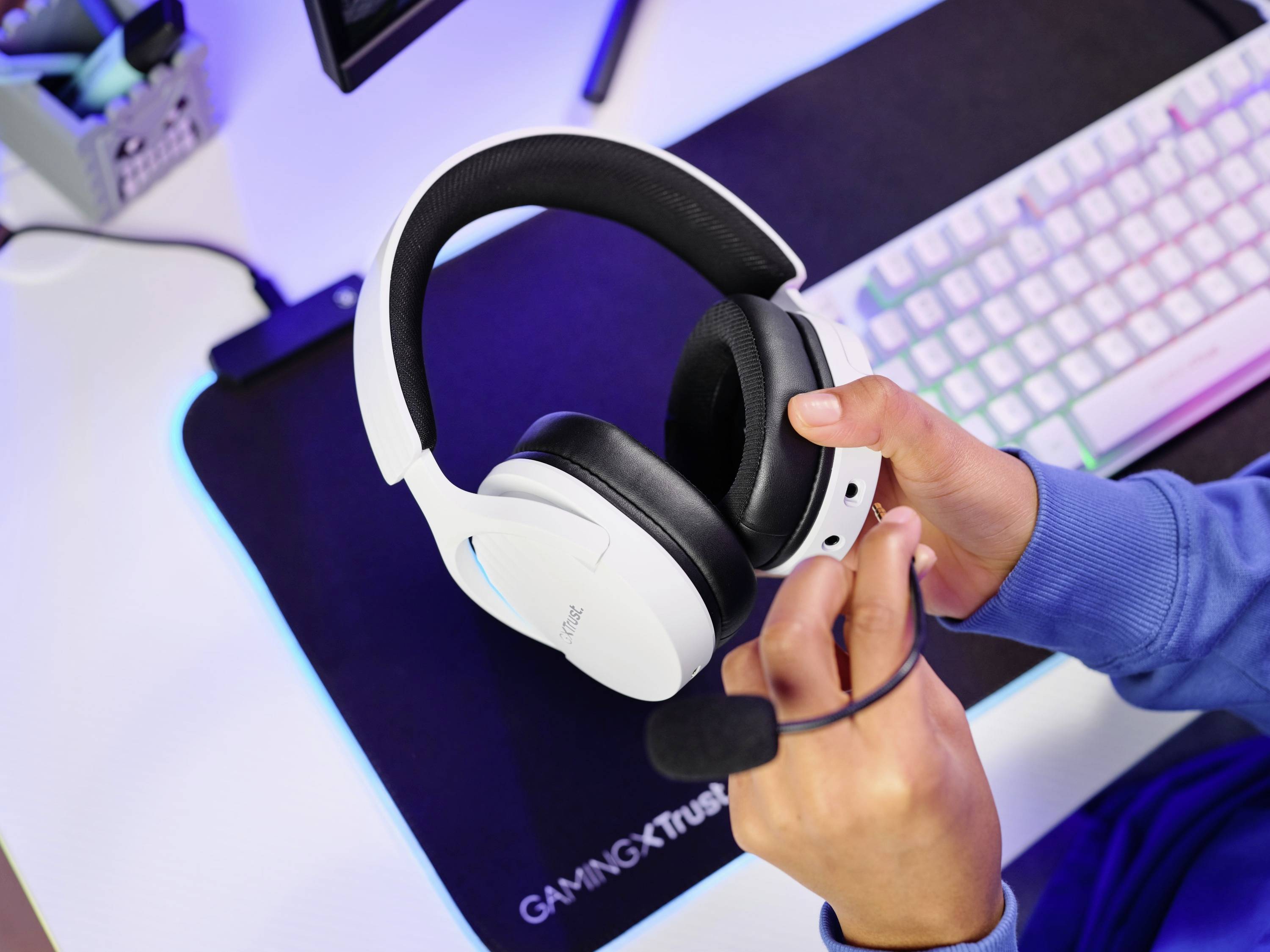 Une personne tient un casque gaming blanc au-dessus d'un bureau avec un clavier et une souris. Un écran est visible en arrière-plan.