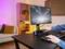Une personne joue à un jeu de course automobile sur un moniteur placé sur un bureau avec un clavier et un éclairage coloré. Un salon est visible en arrière-plan.
