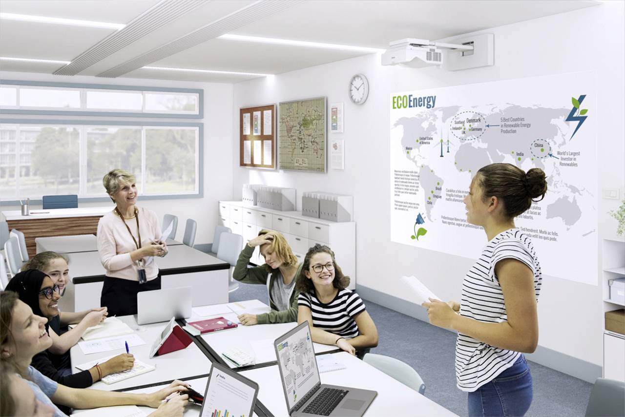 Un groupe d'élèves est assis dans une salle de classe ; une personne est debout et présente une infographie sur 'ECO Energy' au mur.