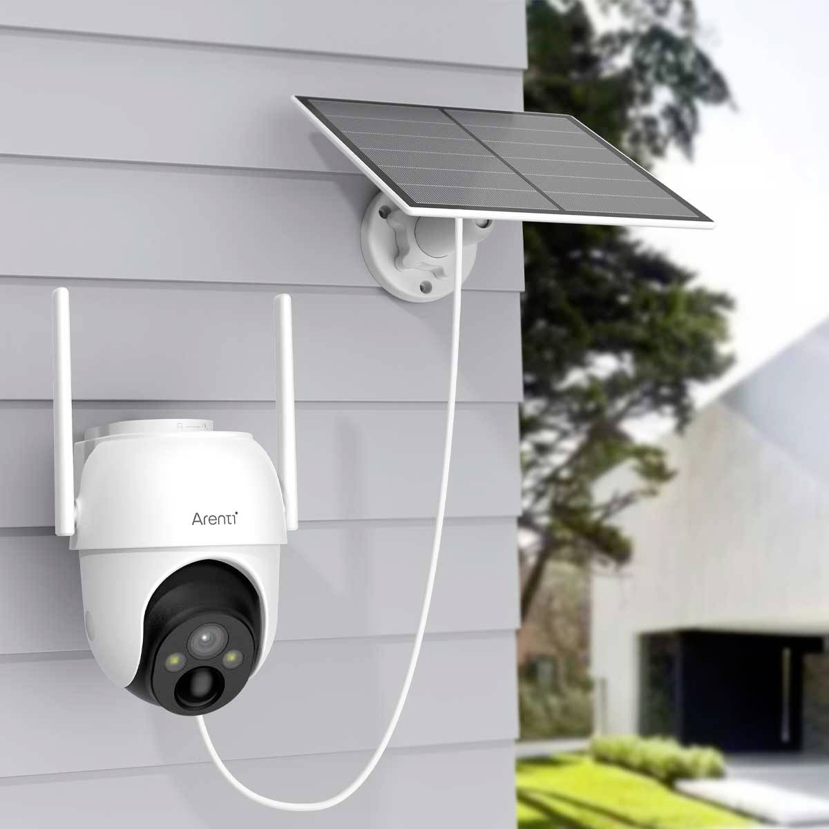 Caméra de surveillance équipée d'un panneau solaire, installée sur un mur de maison ; la caméra possède deux antennes et est protégée contre les intempéries. Un jardin est visible en arrière-plan.