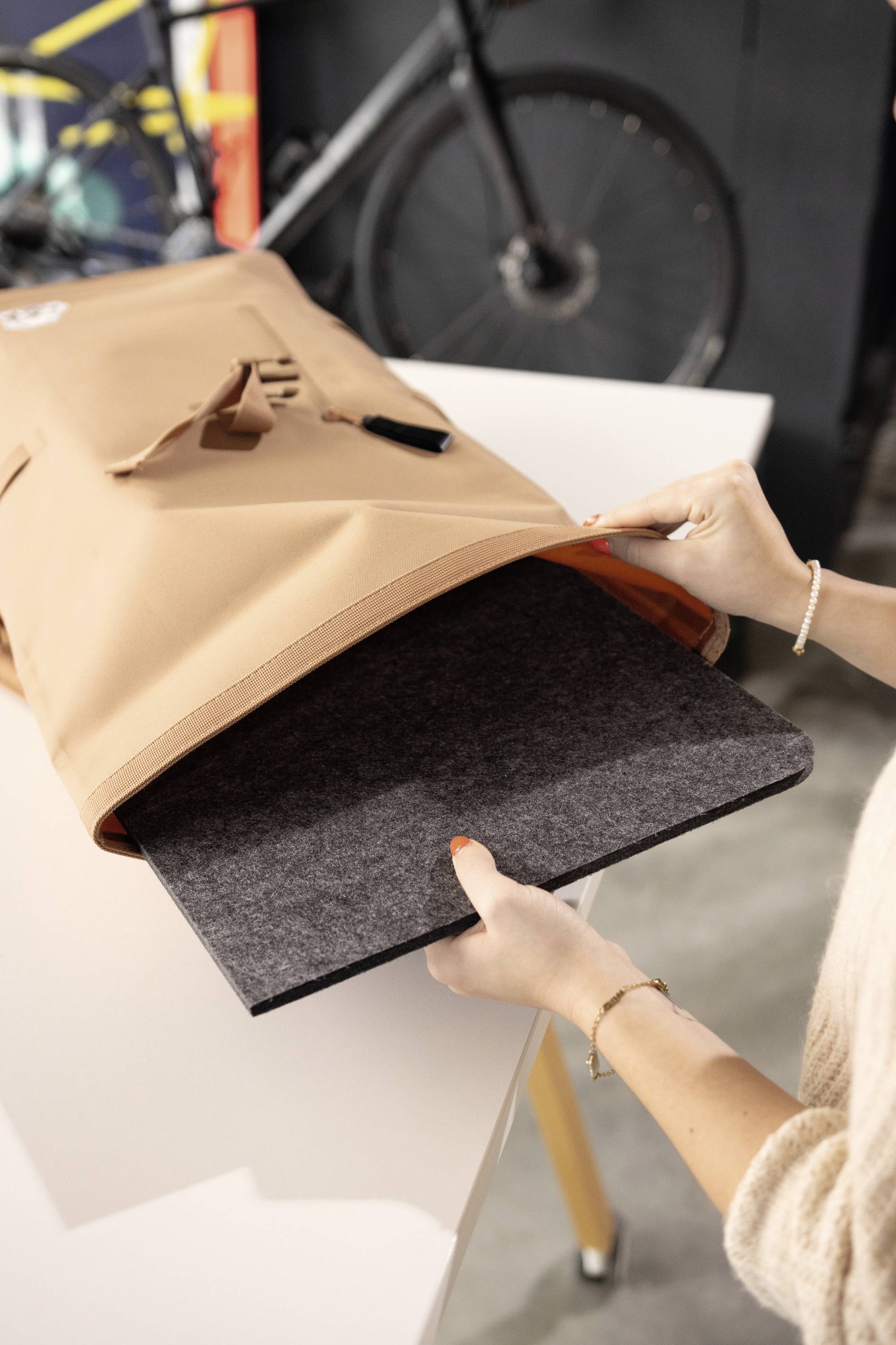 Une personne range un ordinateur portable dans un sac beige posé sur une table. Un vélo est visible en arrière-plan.