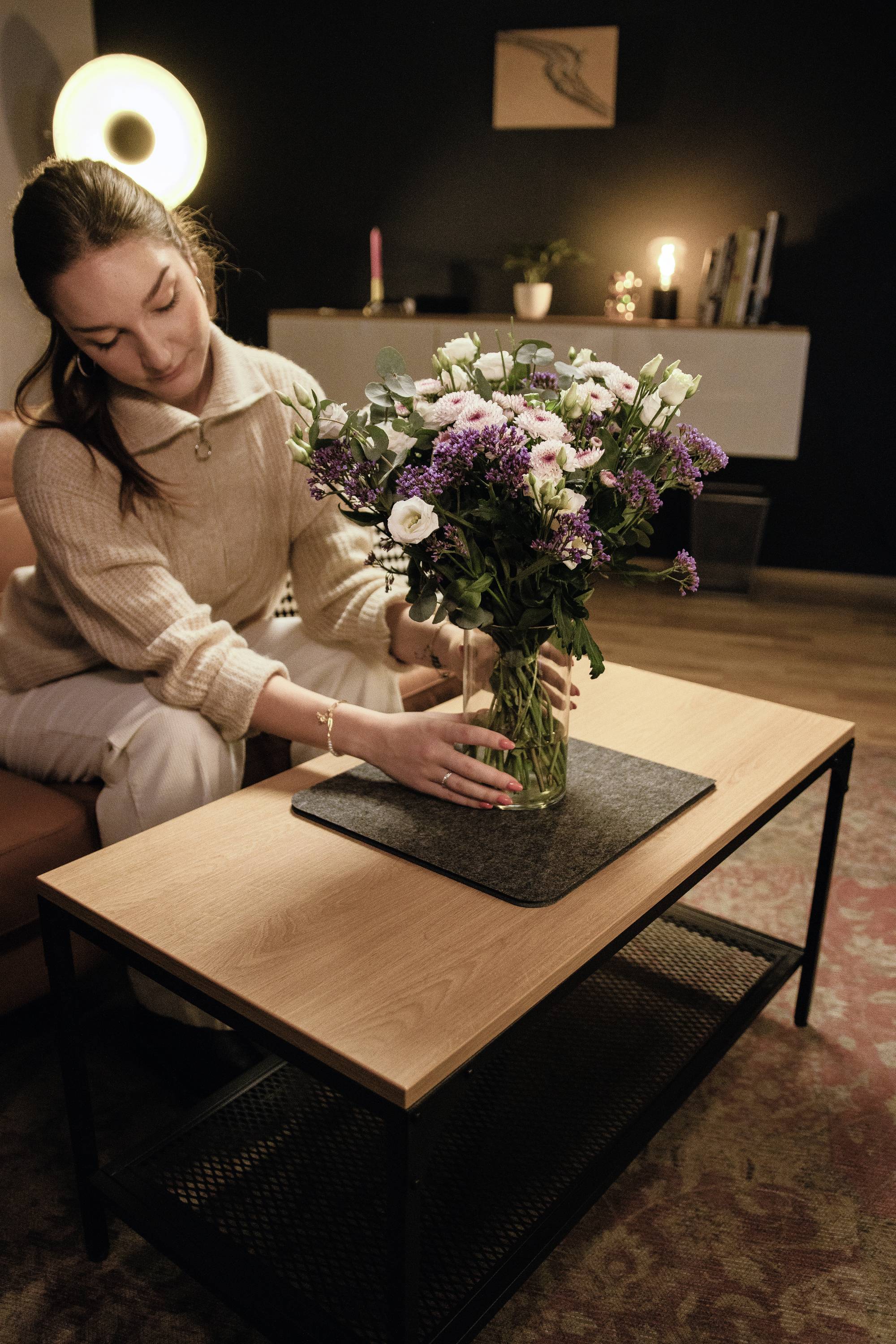 Une femme pose un bouquet de fleurs sur une table de salon. Au fond, on aperçoit une lampe et des décorations.
