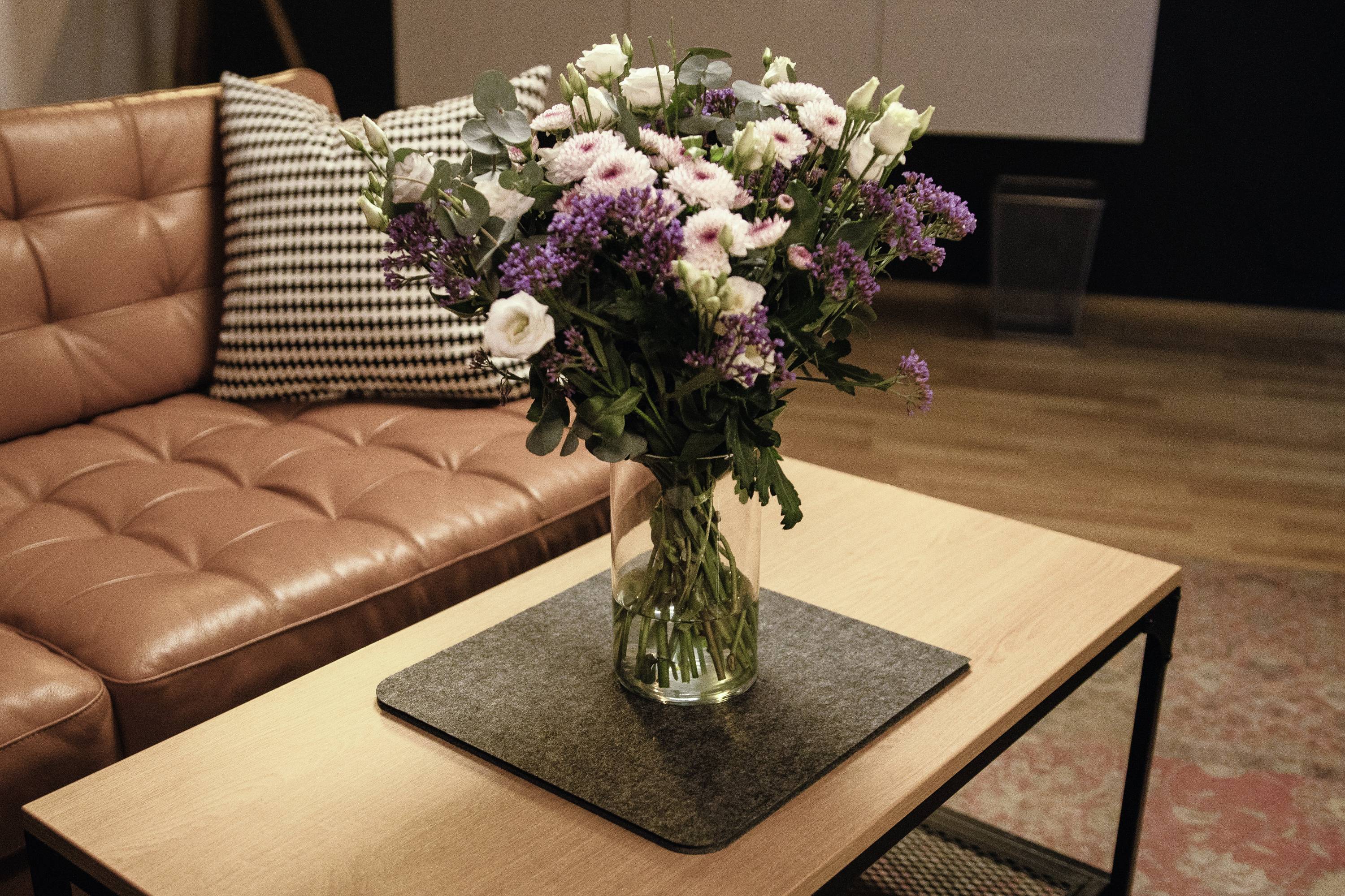 Bouquet de fleurs aux pétales mauves et blancs dans un vase en verre posé sur une table basse, devant un canapé en cuir marron, dans le salon.
