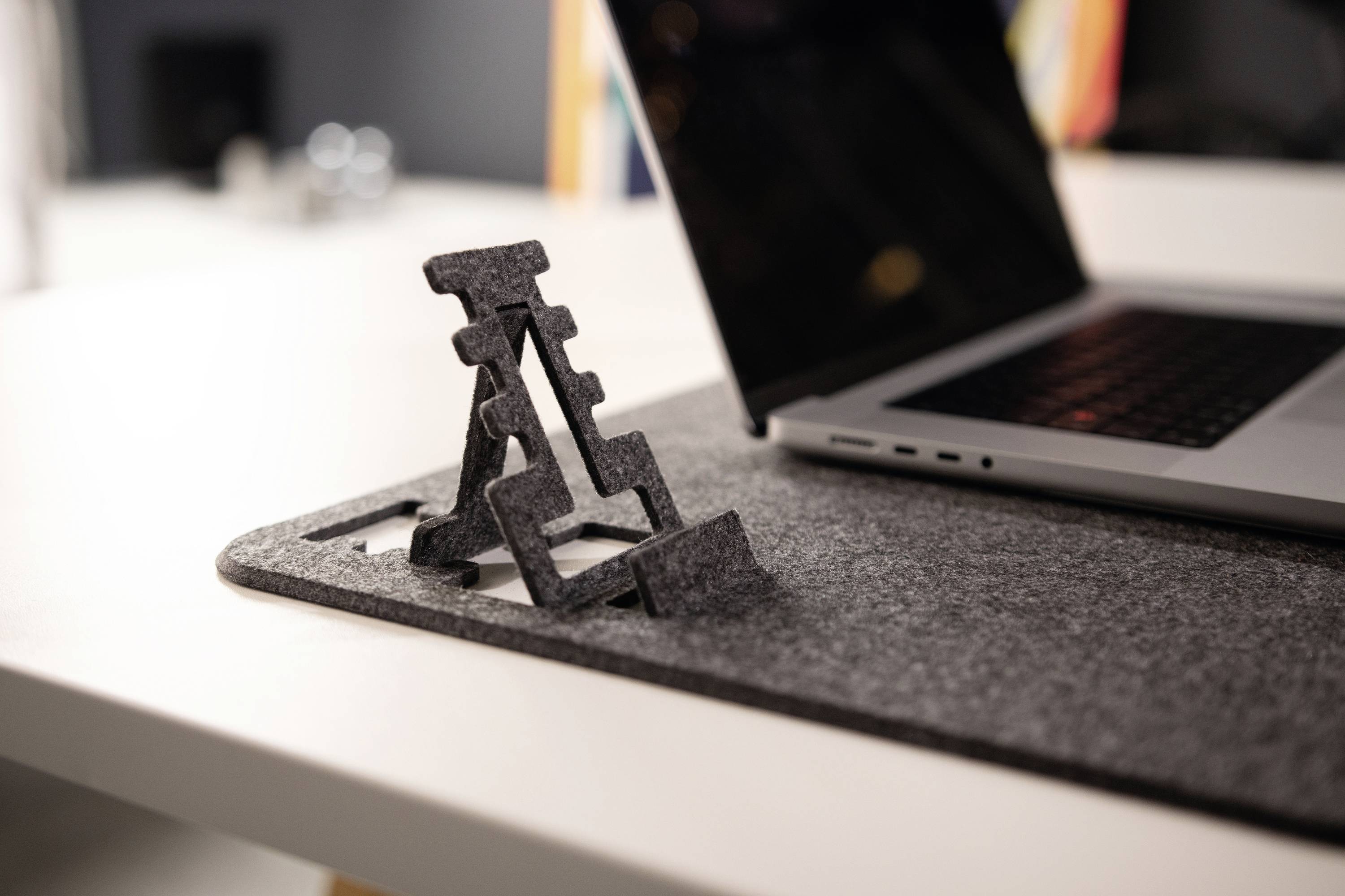 Un ordinateur portable est posé sur un pad en feutre sur une table. À côté de l'ordinateur portable, un support pliable en métal est visible.