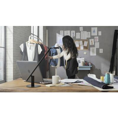 Une designer est au téléphone dans un atelier de mode. Elle examine des vêtements suspendus à un portant. Des croquis sont visibles en arrière-plan.