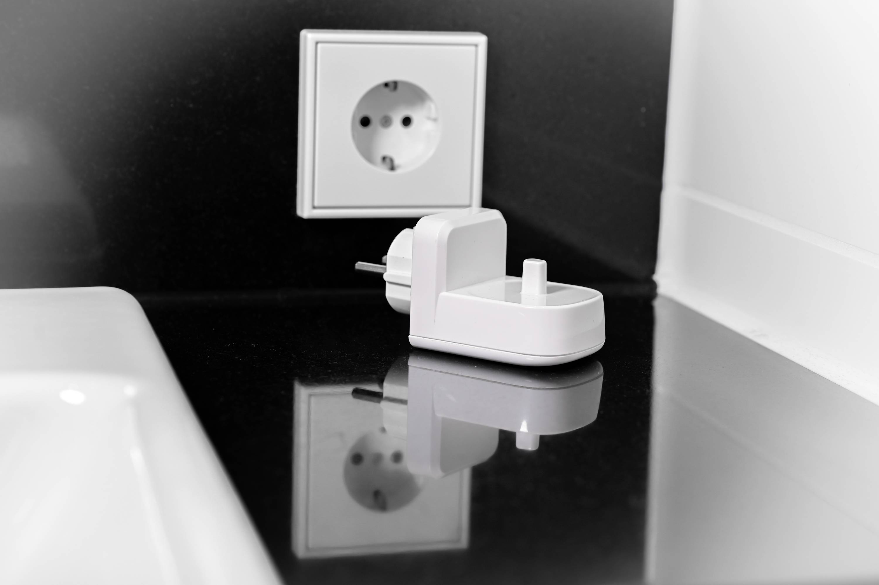 Une prise électrique blanche sur un mur noir avec une fiche secteur blanche posée sur le plan de travail. Il y a un lavabo à côté.