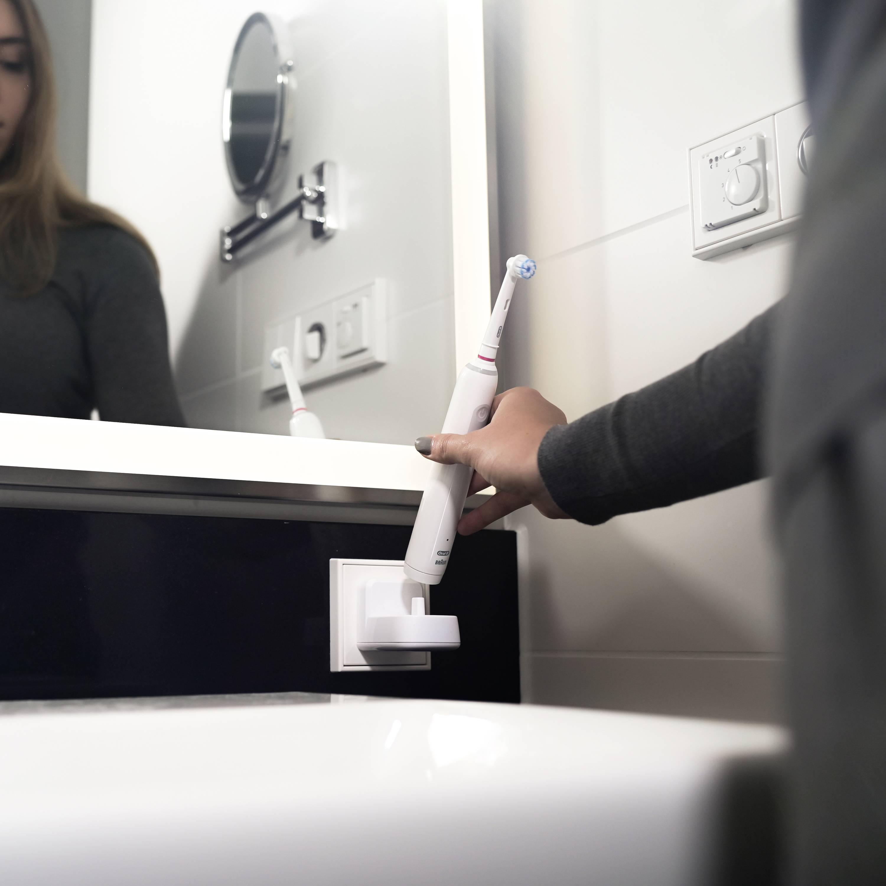 Une personne charge une brosse à dents électrique à une prise électrique dans la salle de bain. Un miroir et un interrupteur sont visibles en arrière-plan.