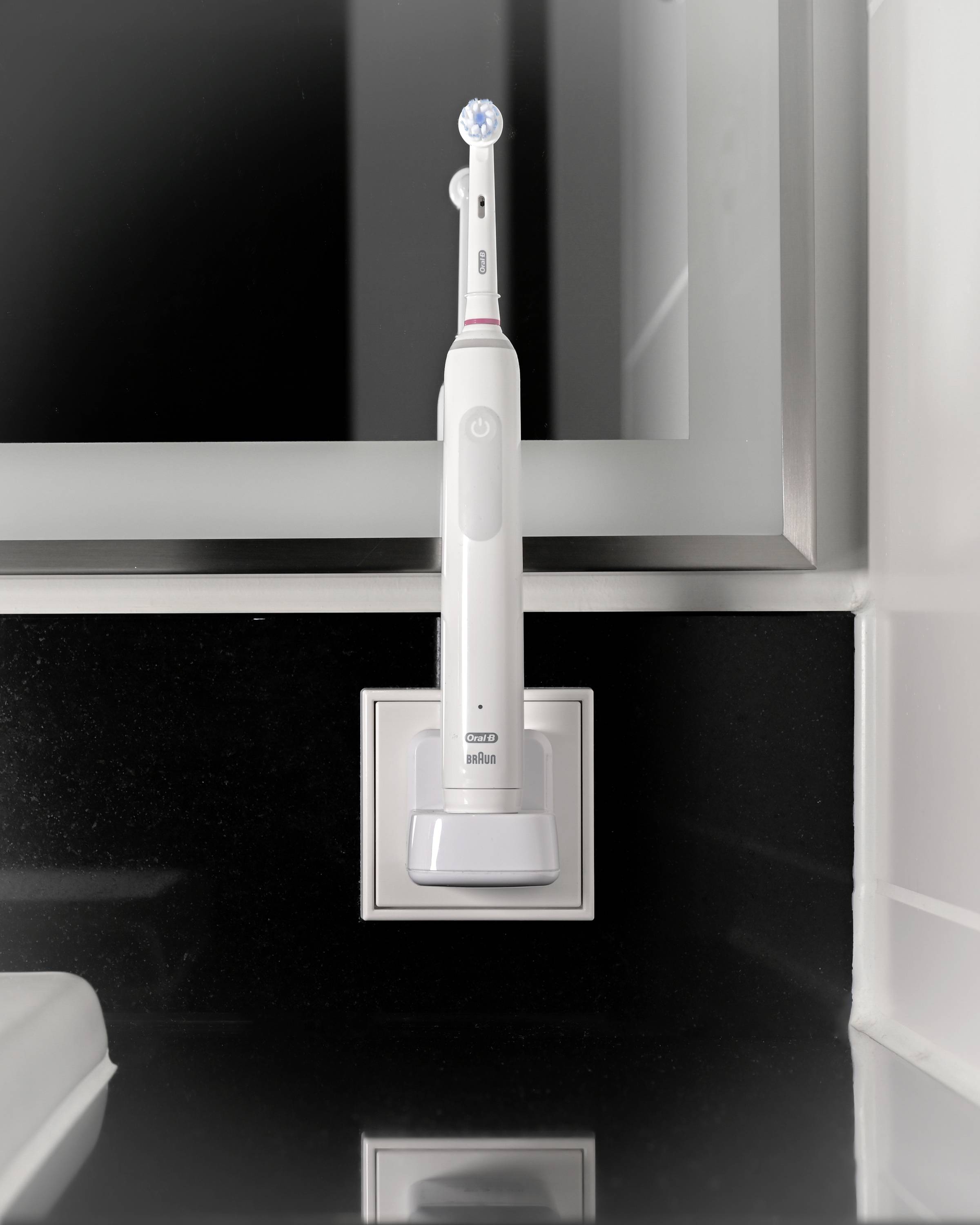 Brosse à dents électrique dans sa station de charge posée sur un support noir, montée devant un miroir argenté dans la salle de bain.