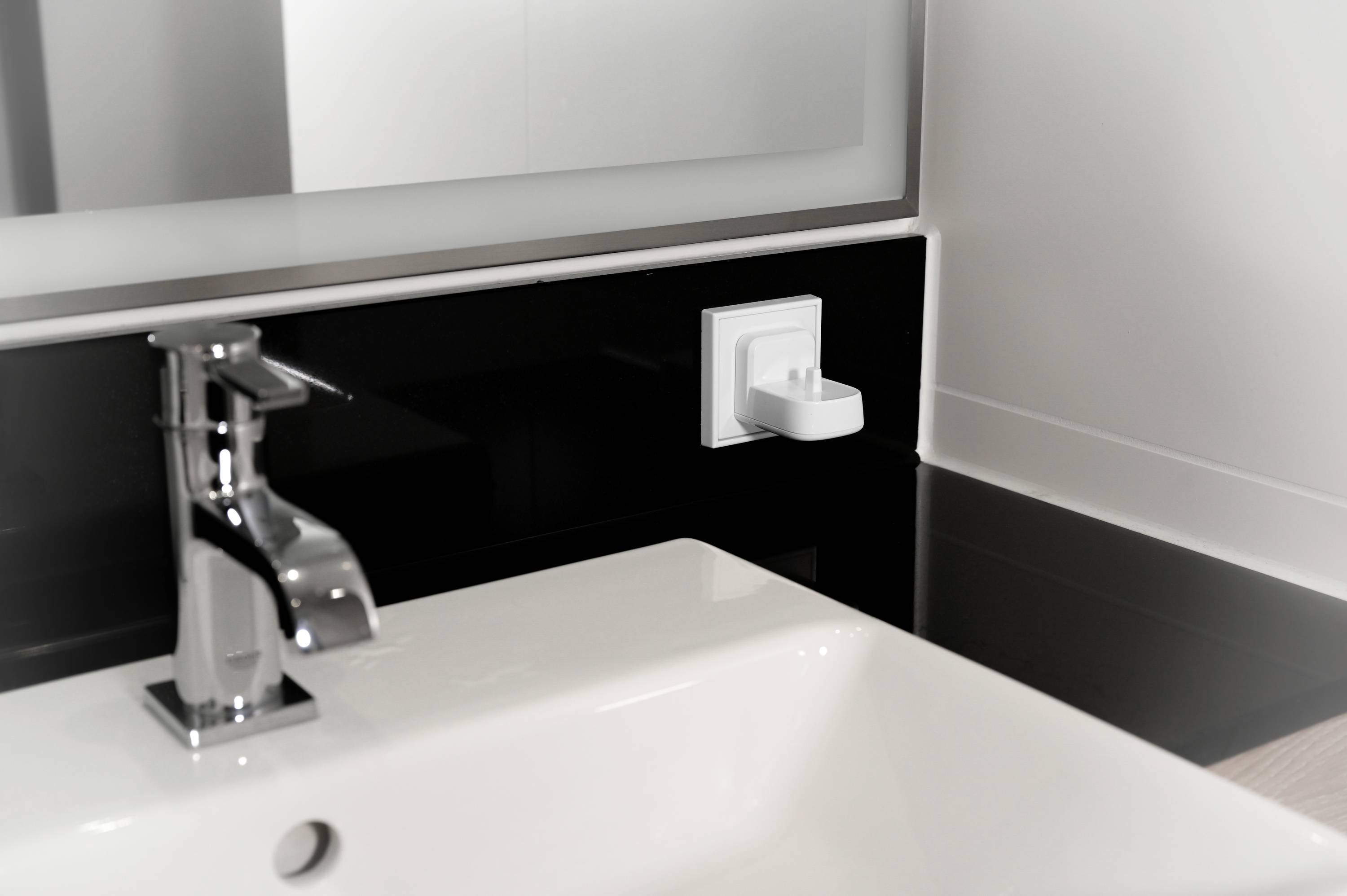 Un lavabo avec un robinet moderne, au-dessous un panneau noir. Un adaptateur électrique blanc est monté sur le mur.