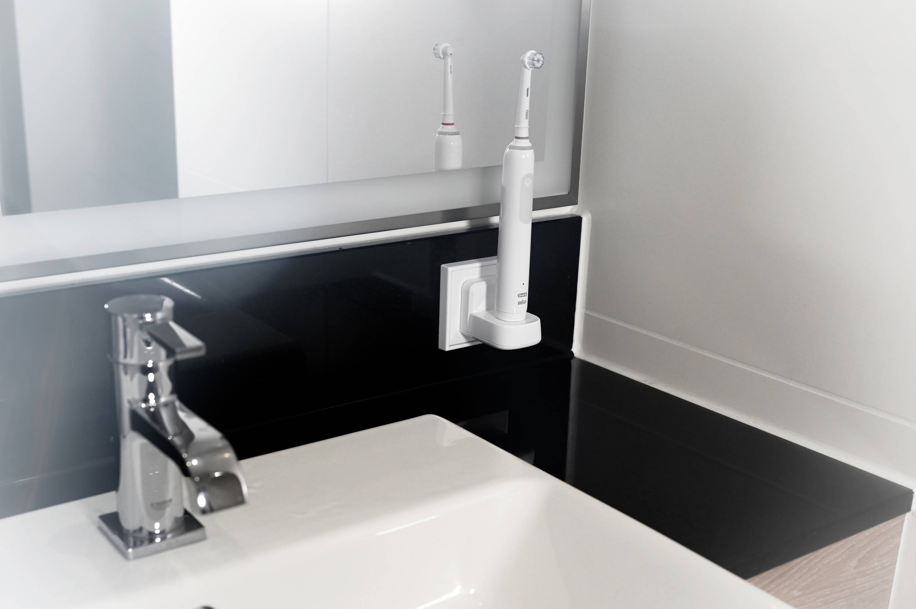 Une salle de bain avec un lavabo blanc et un robinet, à côté se trouve une brosse à dents électrique dans un support fixé au mur.