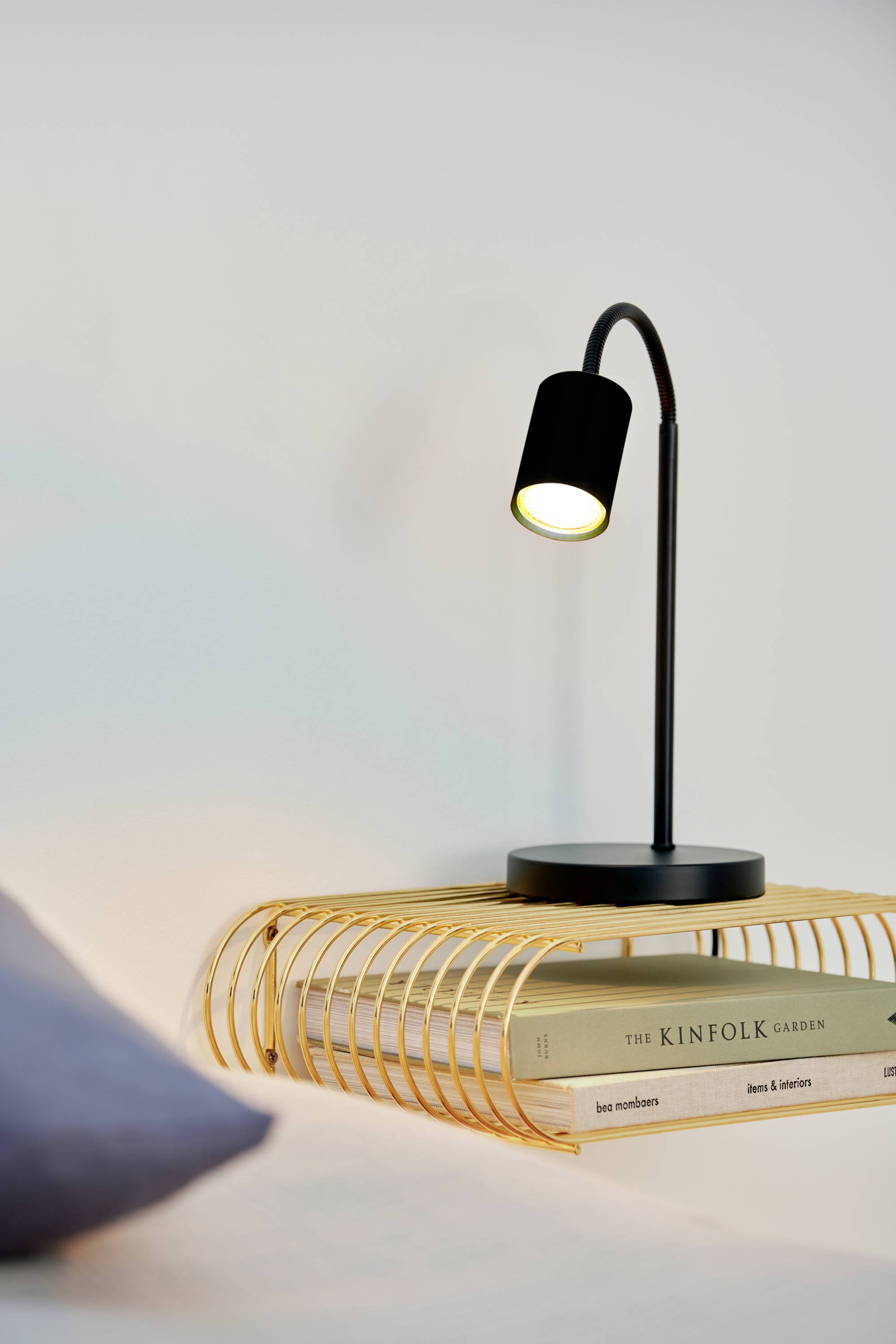Une lampe de table moderne à col incliné éclaire trois livres sur une étagère élégante et dorée.