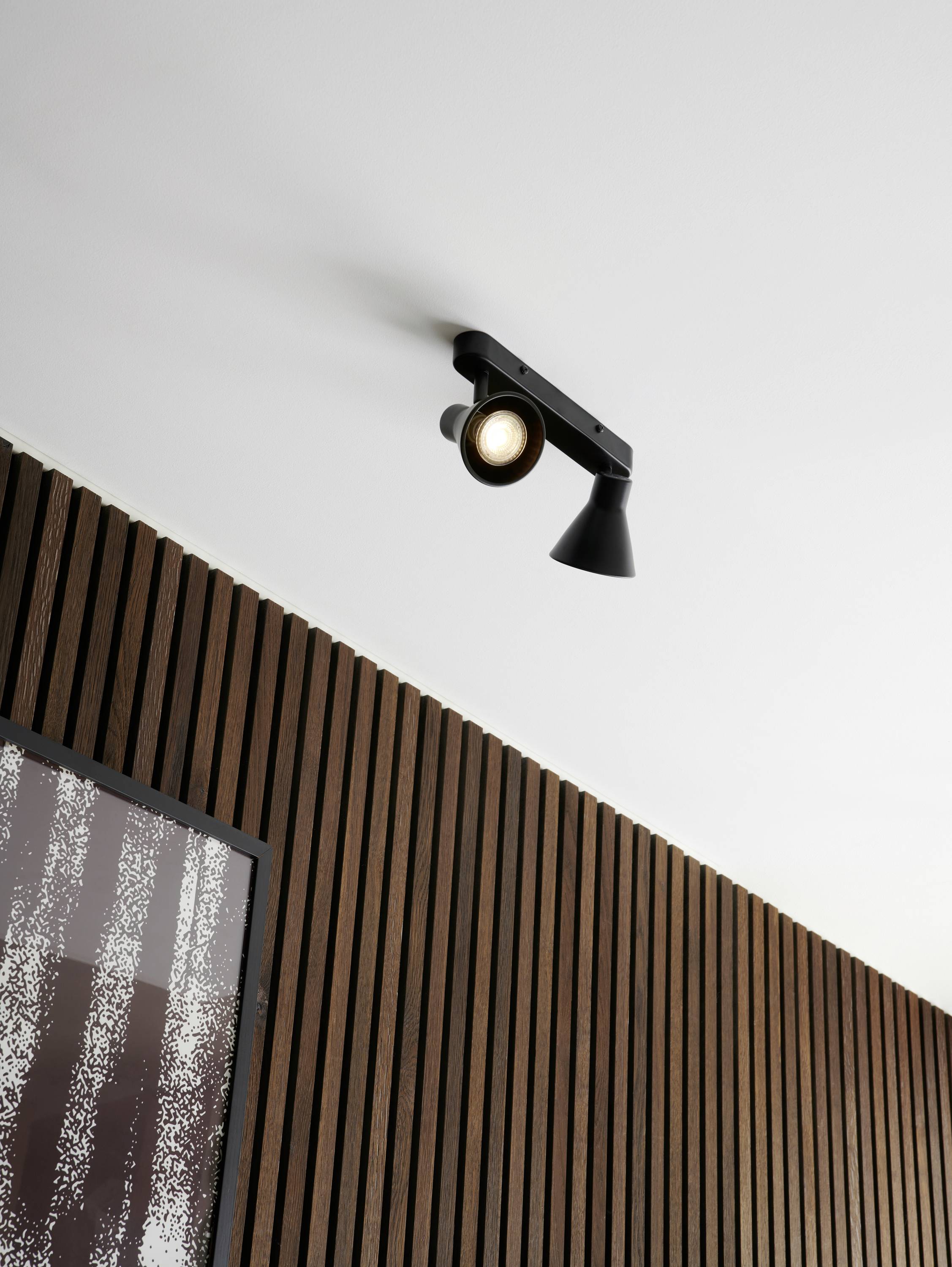 Un projecteur noir au design moderne, fixé au plafond d'une pièce, illumine un revêtement mural en bois à côté d'un tableau partiellement visible.
