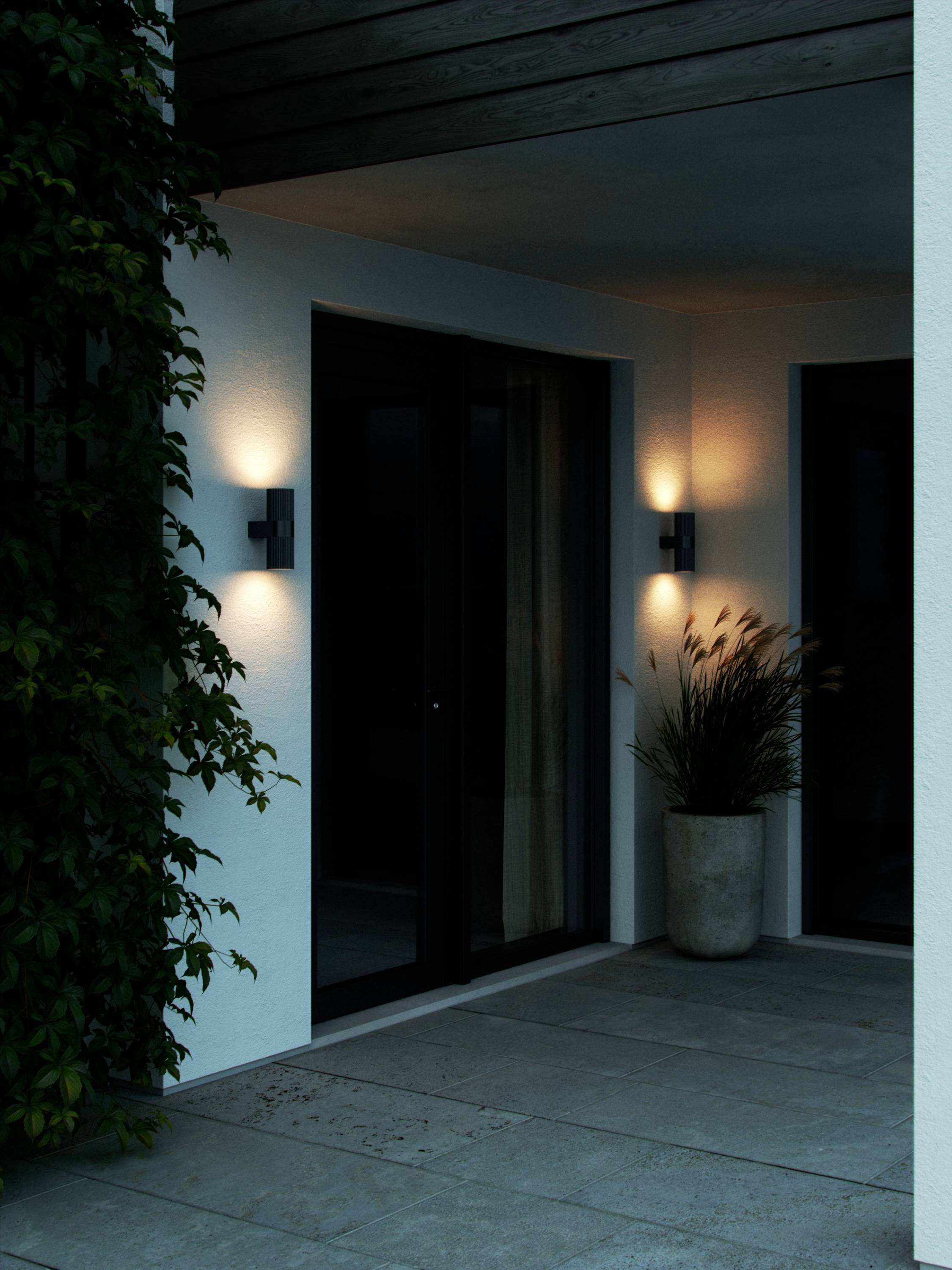 Entrée de maison éclairée avec façade vitrée, lampe murale moderne et plante en pot ; atmosphère paisible et accueillante en soirée.