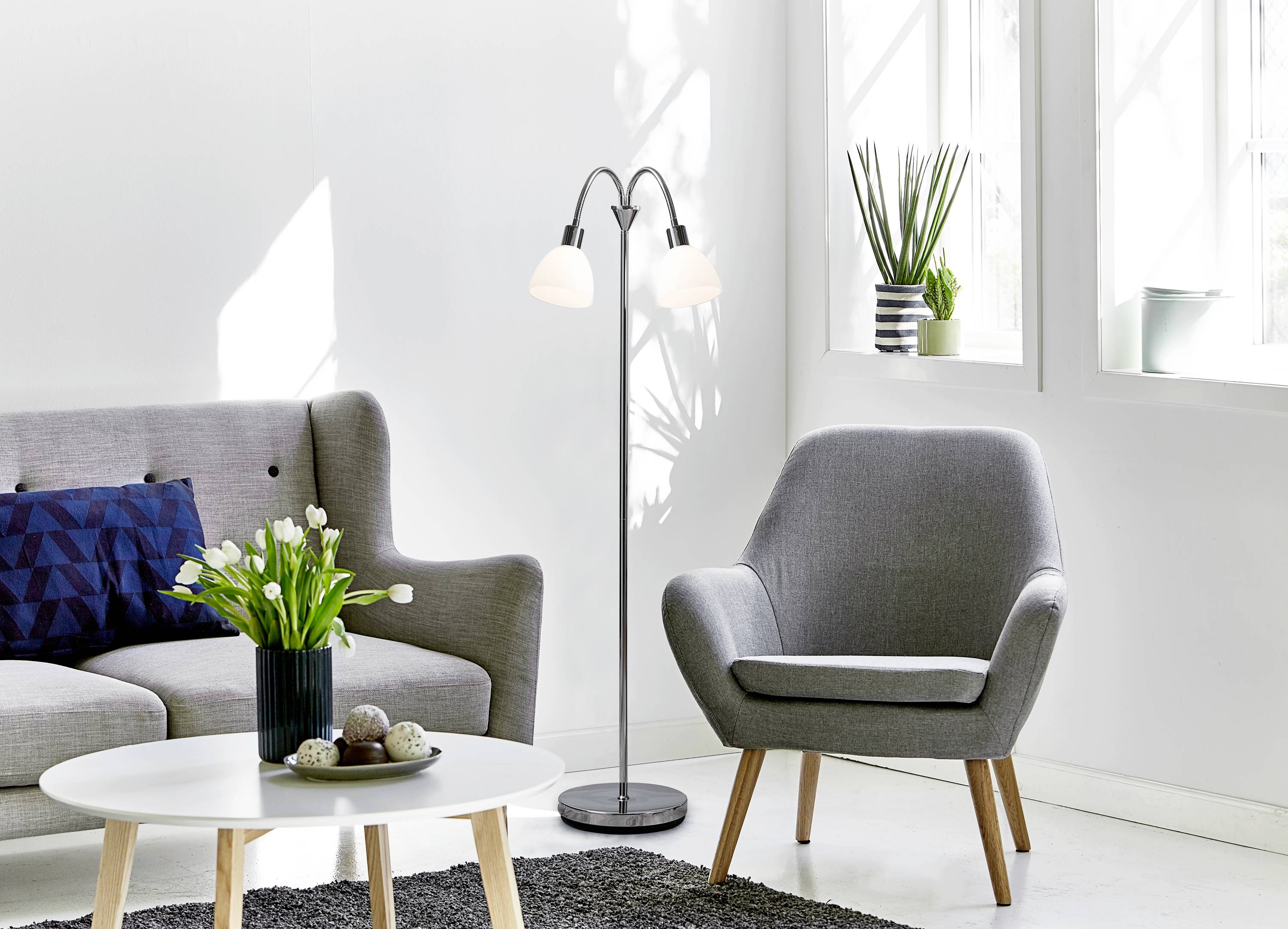 Salon lumineux de style scandinave avec canapé et fauteuil gris, lampadaire, murs blancs, table, tapis et plantes.