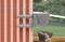 L'image montre un mur de briques avec des poutres en acier apparentes à gauche et un balcon avec des meubles en rotin à droite, avec des arbres verts en arrière-plan.