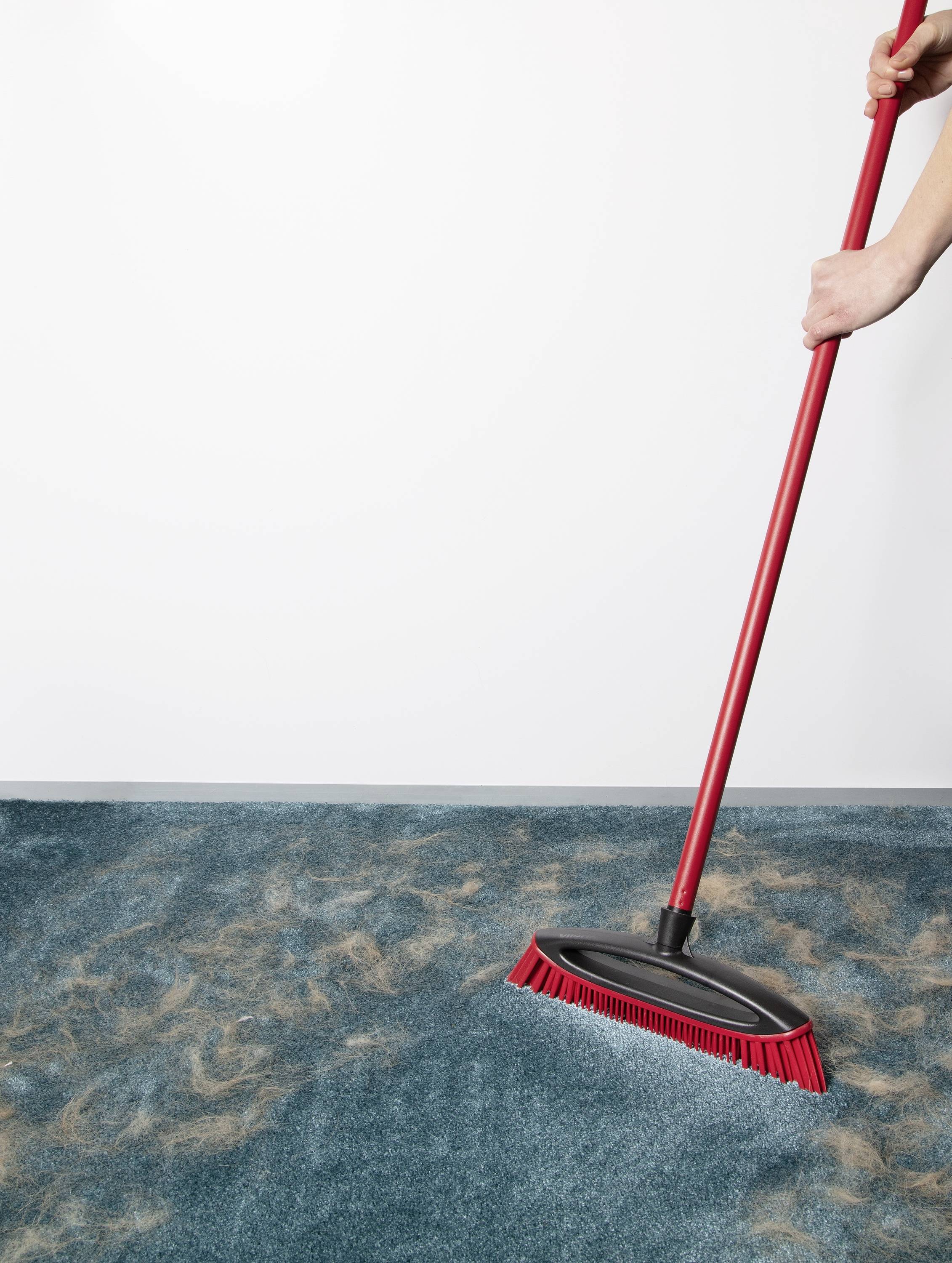 Une main nettoie des cheveux d'un tapis bleu avec un balai rouge.