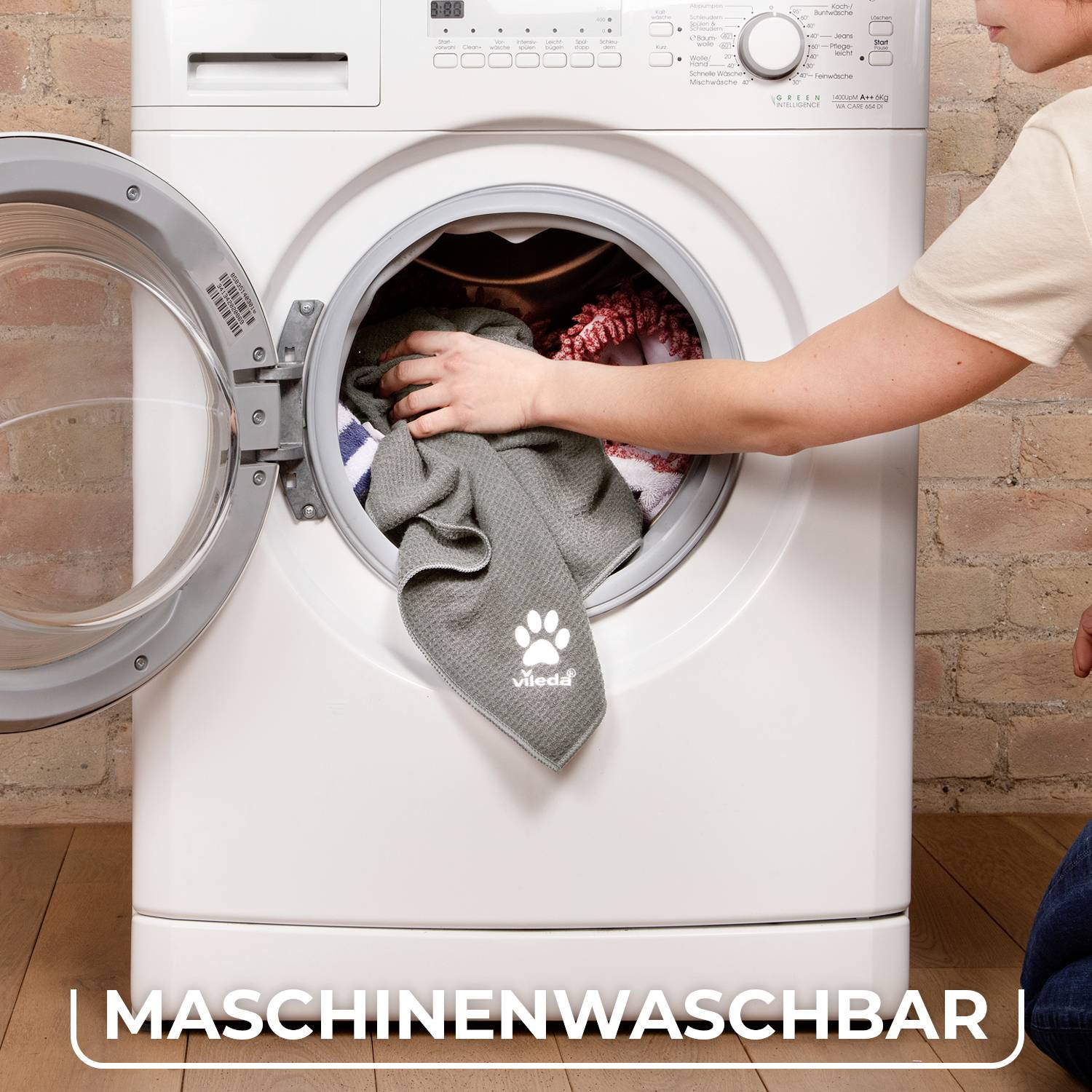 Une personne met des vêtements dans une machine à laver ouverte. Sur une serviette grise, il est inscrit 'Lavable en machine'.