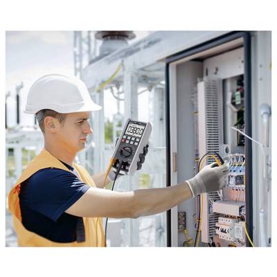 Électricien portant un casque de sécurité testant le câblage dans un panneau de commande électrique ouvert avec un multimètre numérique.