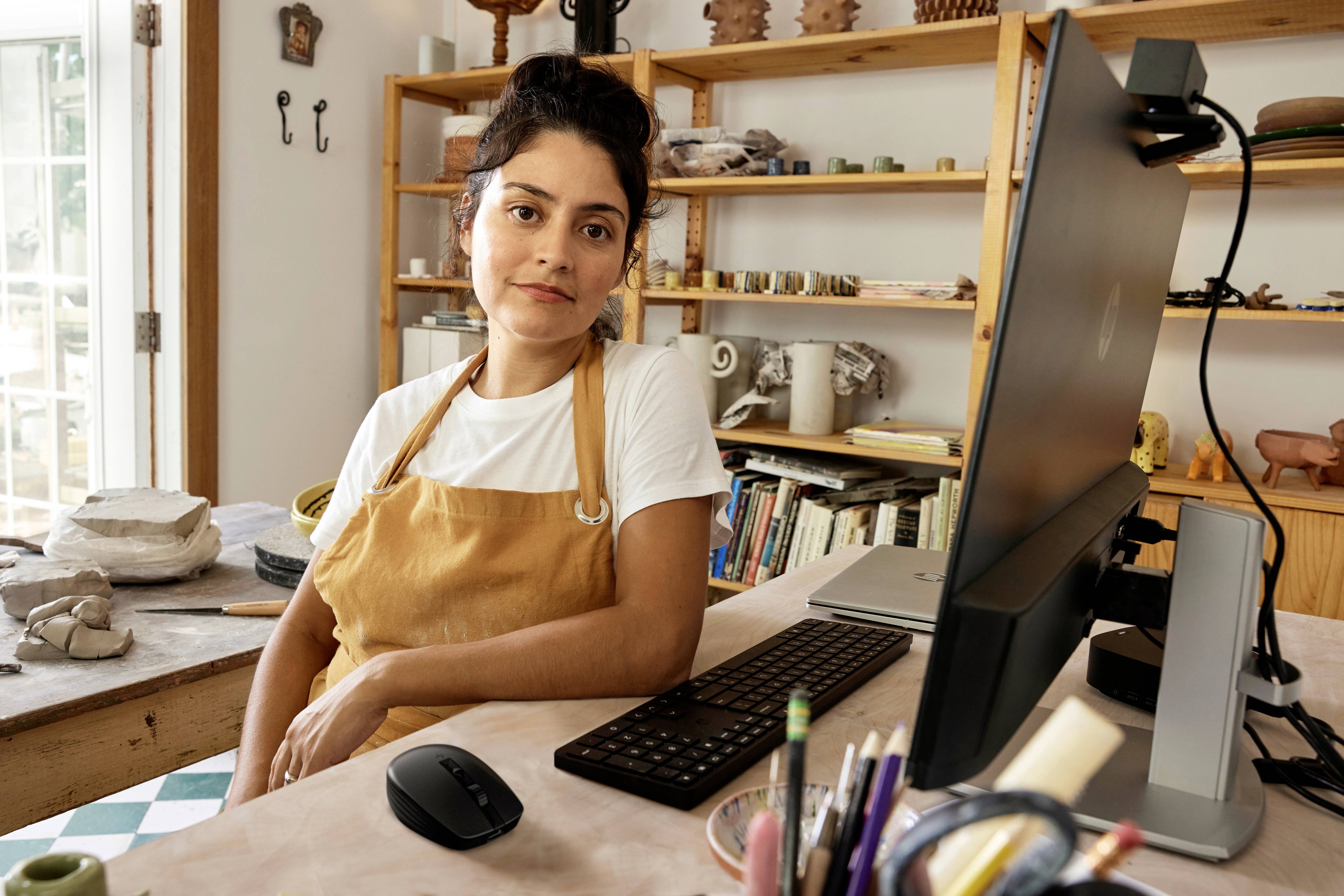 Une personne est assise dans un atelier d'artisanat avec de l'équipement de poterie, porte un tablier et regarde la caméra tout en travaillant sur un ordinateur.