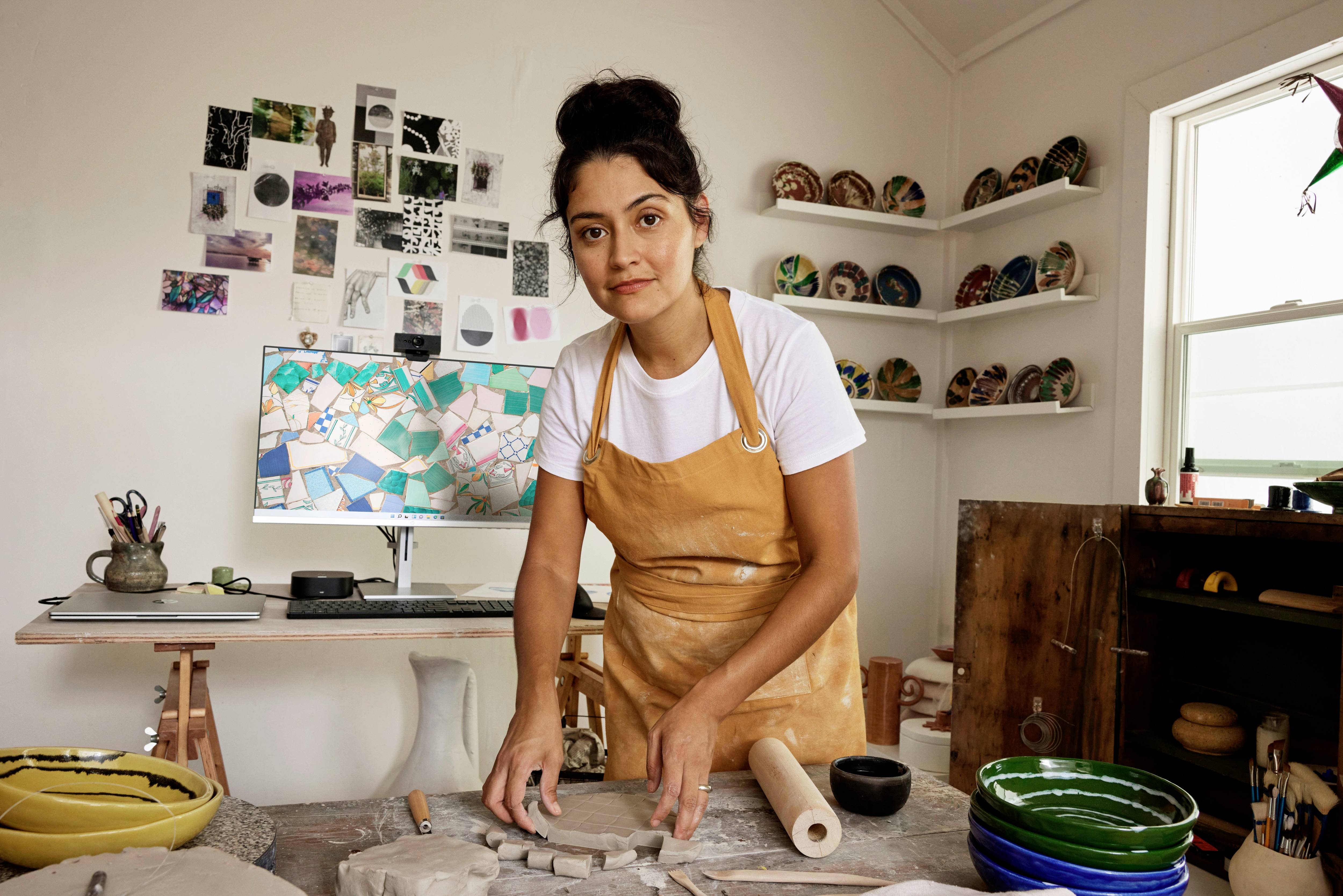 Une femme en tablier travaille sur un projet de céramique dans un atelier ; des pots et des bols l'entourent, des photos au mur servant d'inspiration.