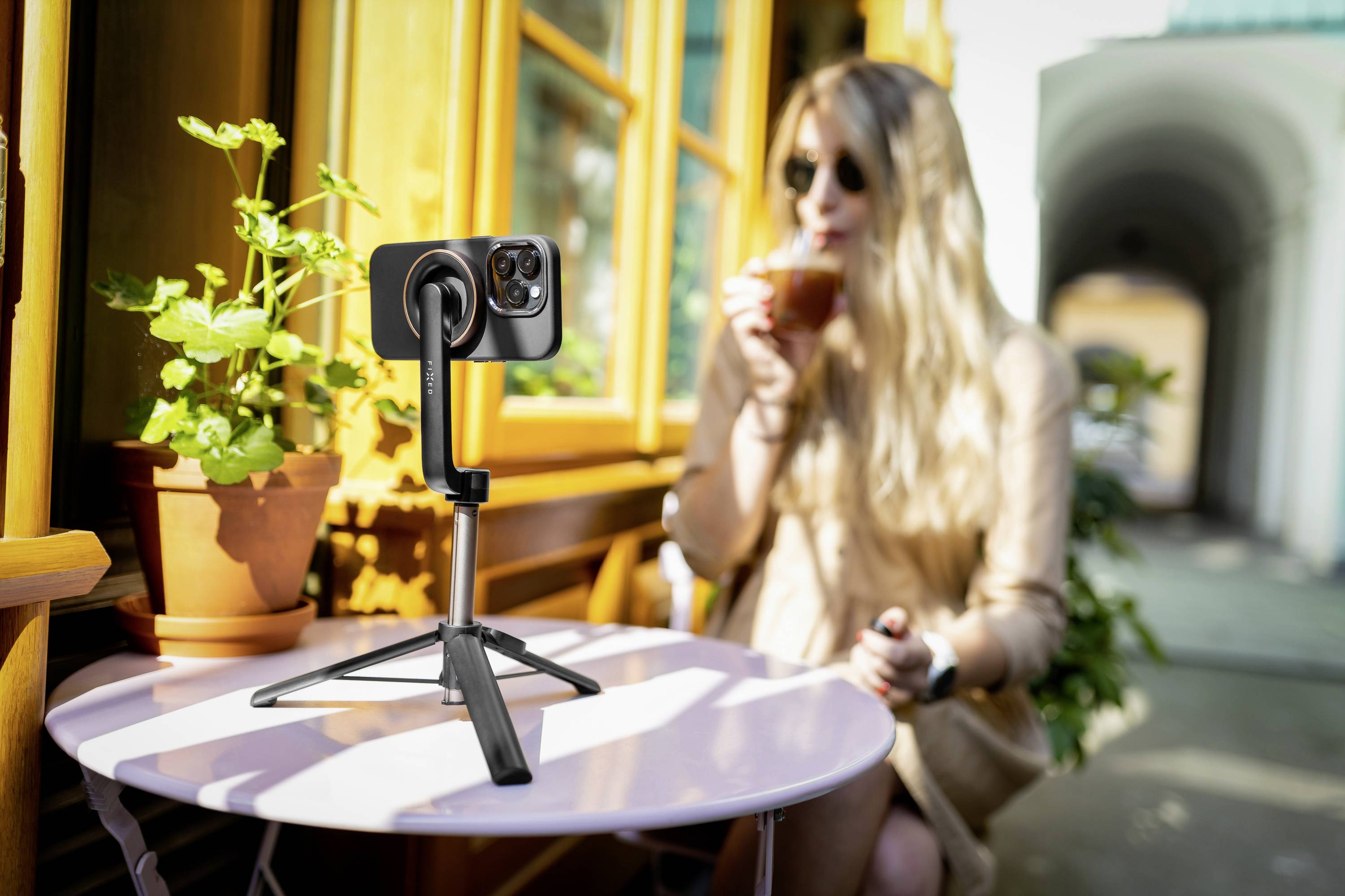 Un smartphone monté sur trépied photographie une femme assise devant un café, buvant un café. La scène transmet une sensation de détente dans un environnement urbain.