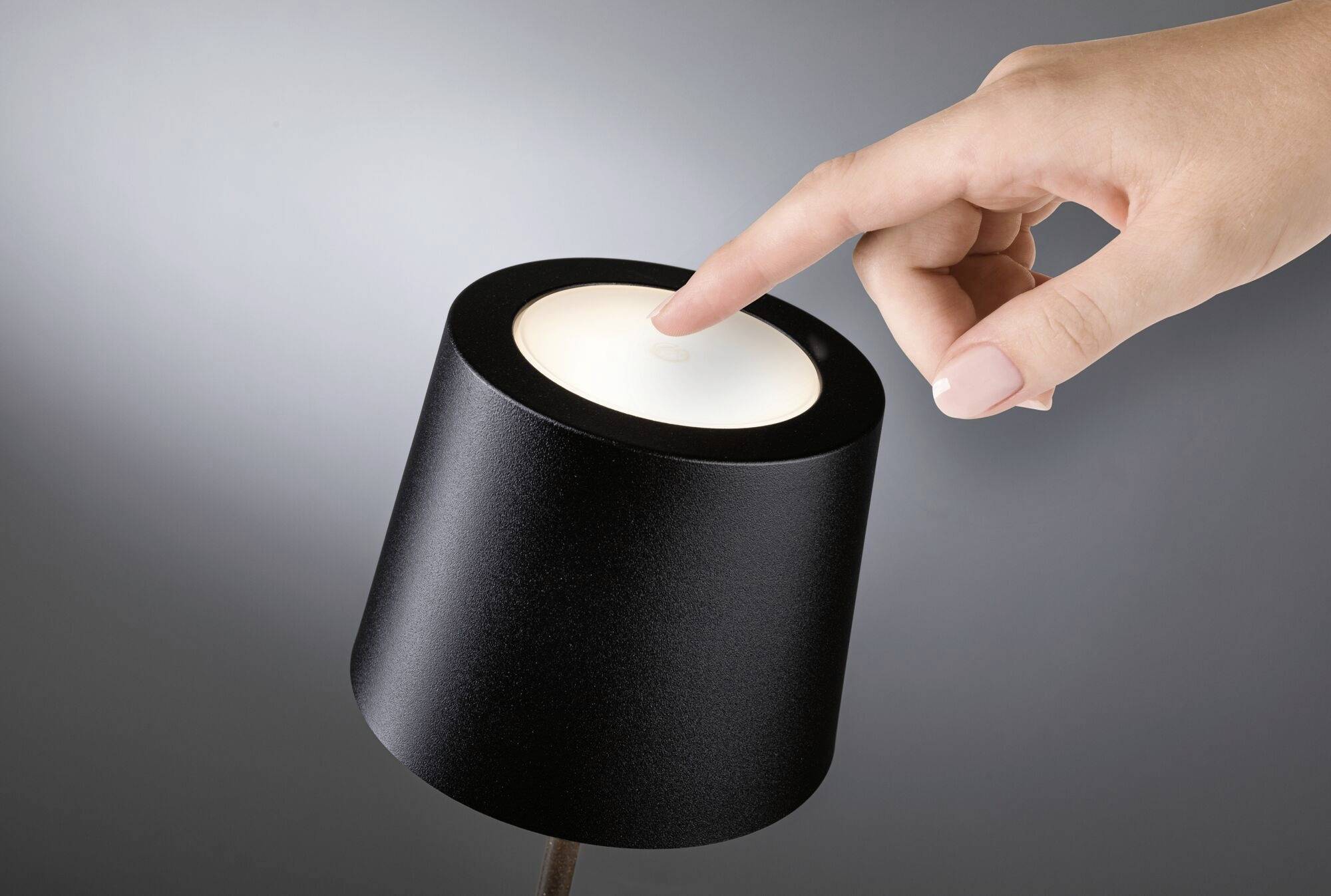 Une main touche le dessus d'une lampe moderne, ronde et noire pour l'allumer, sur un fond gris.