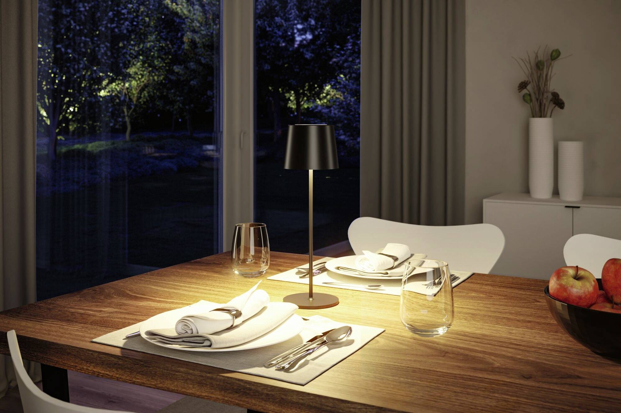 Une table dressée pour deux personnes, chacune avec des couverts, des verres et des serviettes. Une lampe de table éclaire la table. À l'arrière-plan, une fenêtre offre une vue sur un jardin nocturne.