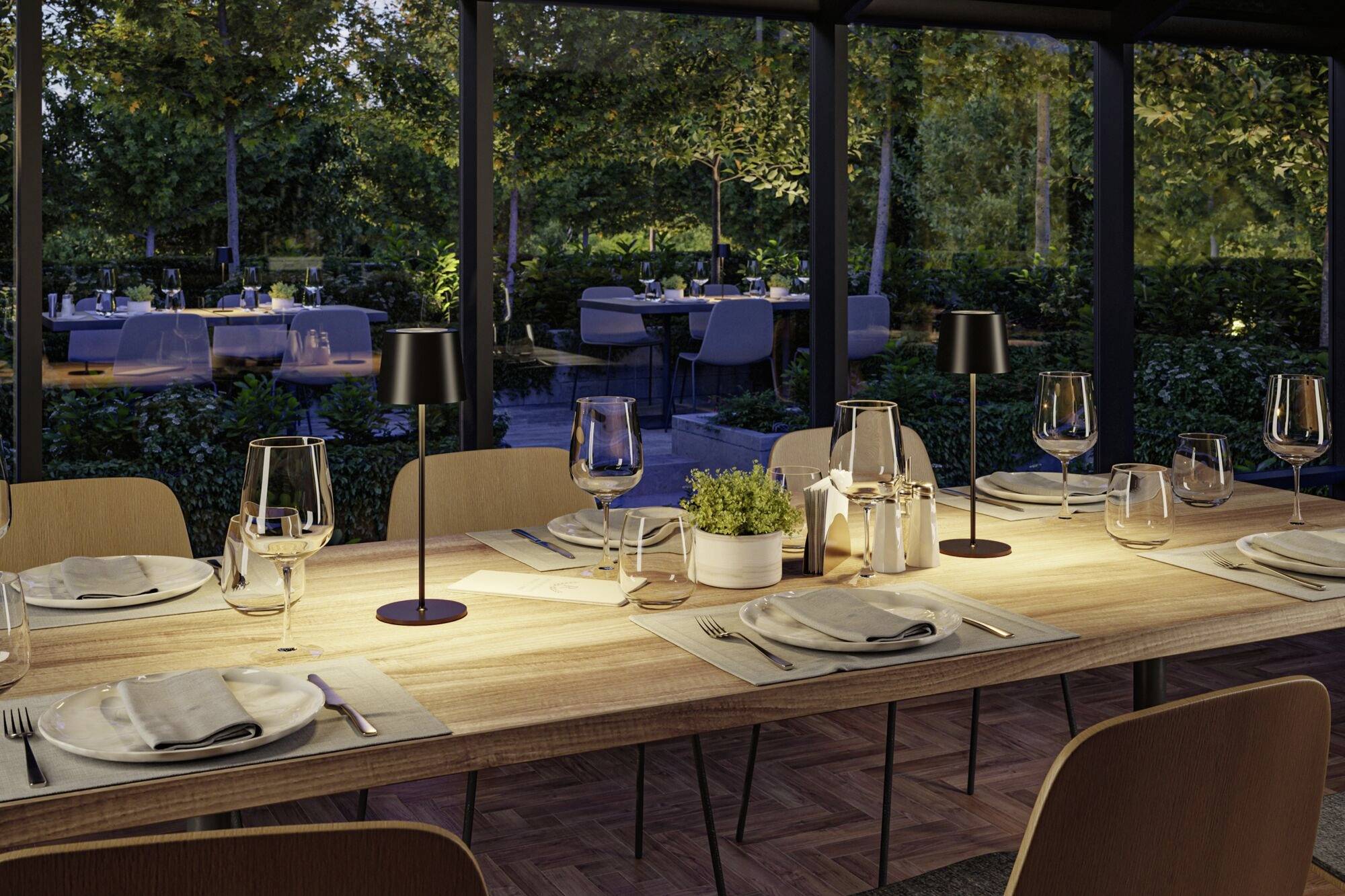 Une table de restaurant élégamment dressée en extérieur, avec des verres à vin, des assiettes et des couverts ; entourée d'aménagements paysagers soignés en fin de journée.
