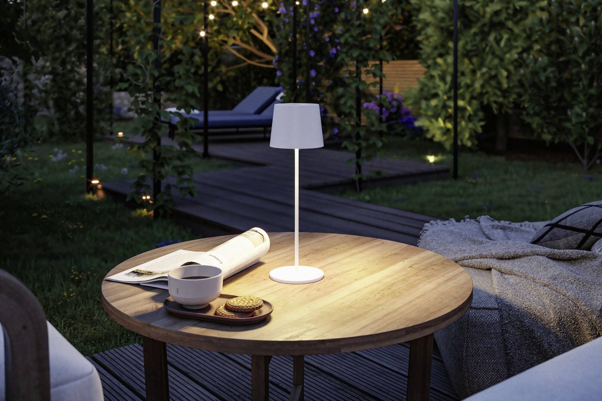 Une table ronde en bois dans le jardin avec une lampe de table blanche, un livre ouvert, une tasse et des cookies. En arrière-plan, un espace de jardin éclairé.