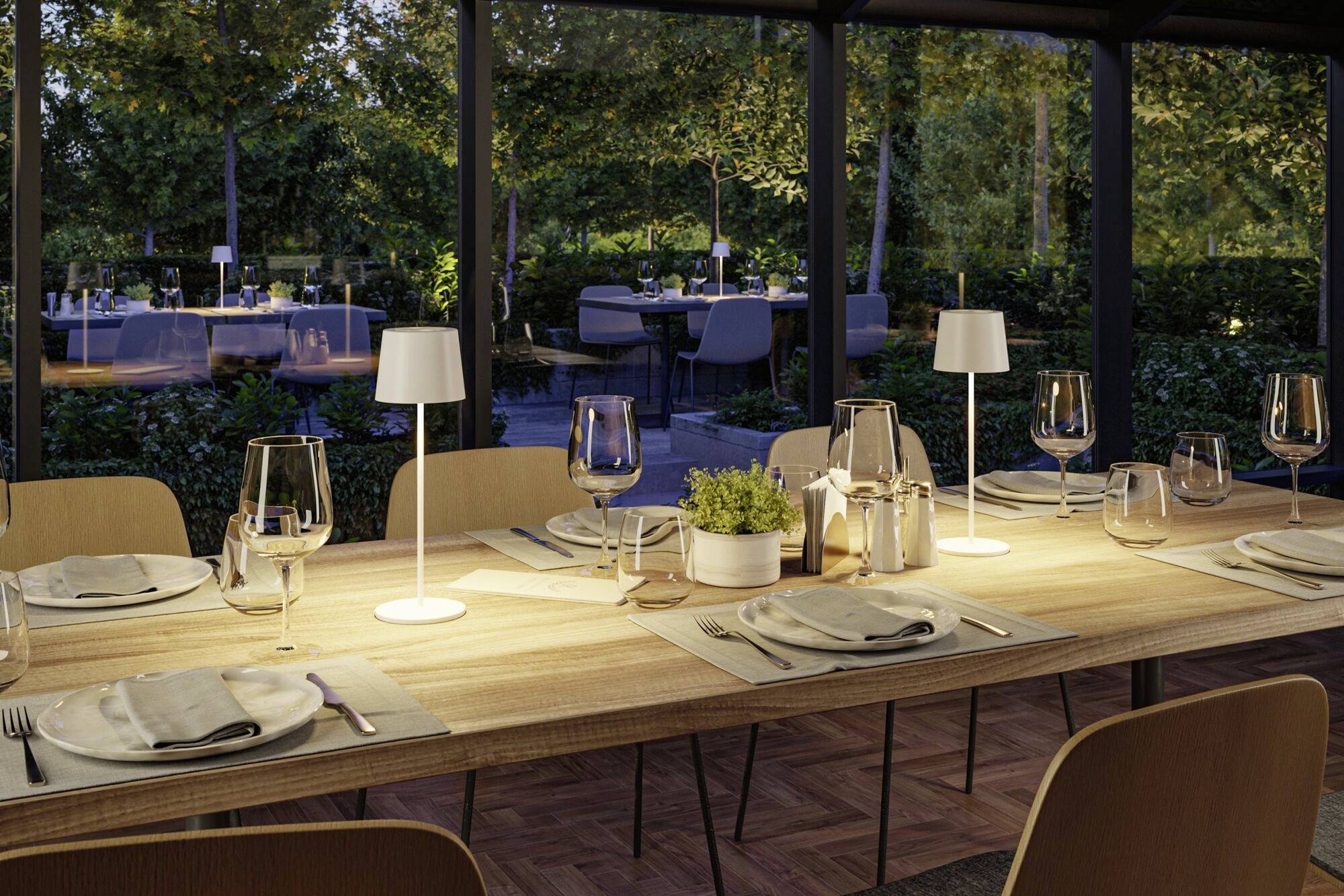 Une table dressée avec élégance dans un restaurant, avec des lampes modernes, des verres et des couverts. À l'extérieur, des arbres et une terrasse sont visibles.