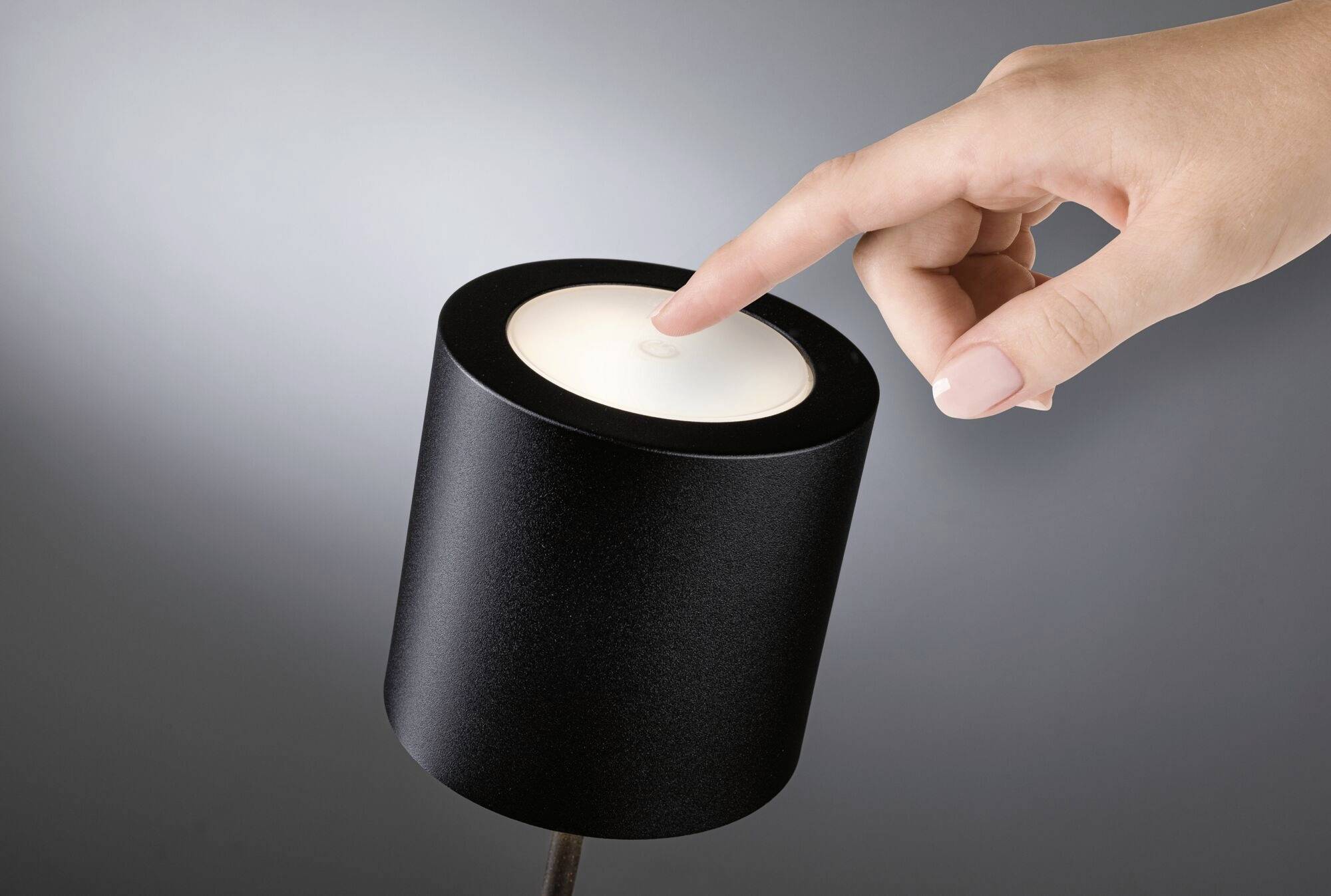 Une main touche le dessus d'une lampe cylindrique noire pour l'allumer ou l'éteindre.
