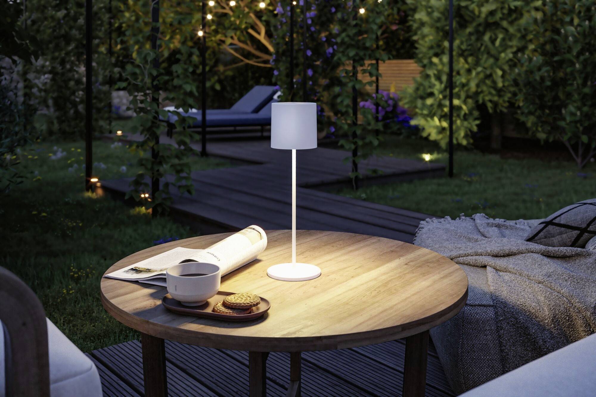 Une table ronde en bois dans le jardin au crépuscule, avec une petite lampe, une tasse, des biscuits et un livre ouvert.