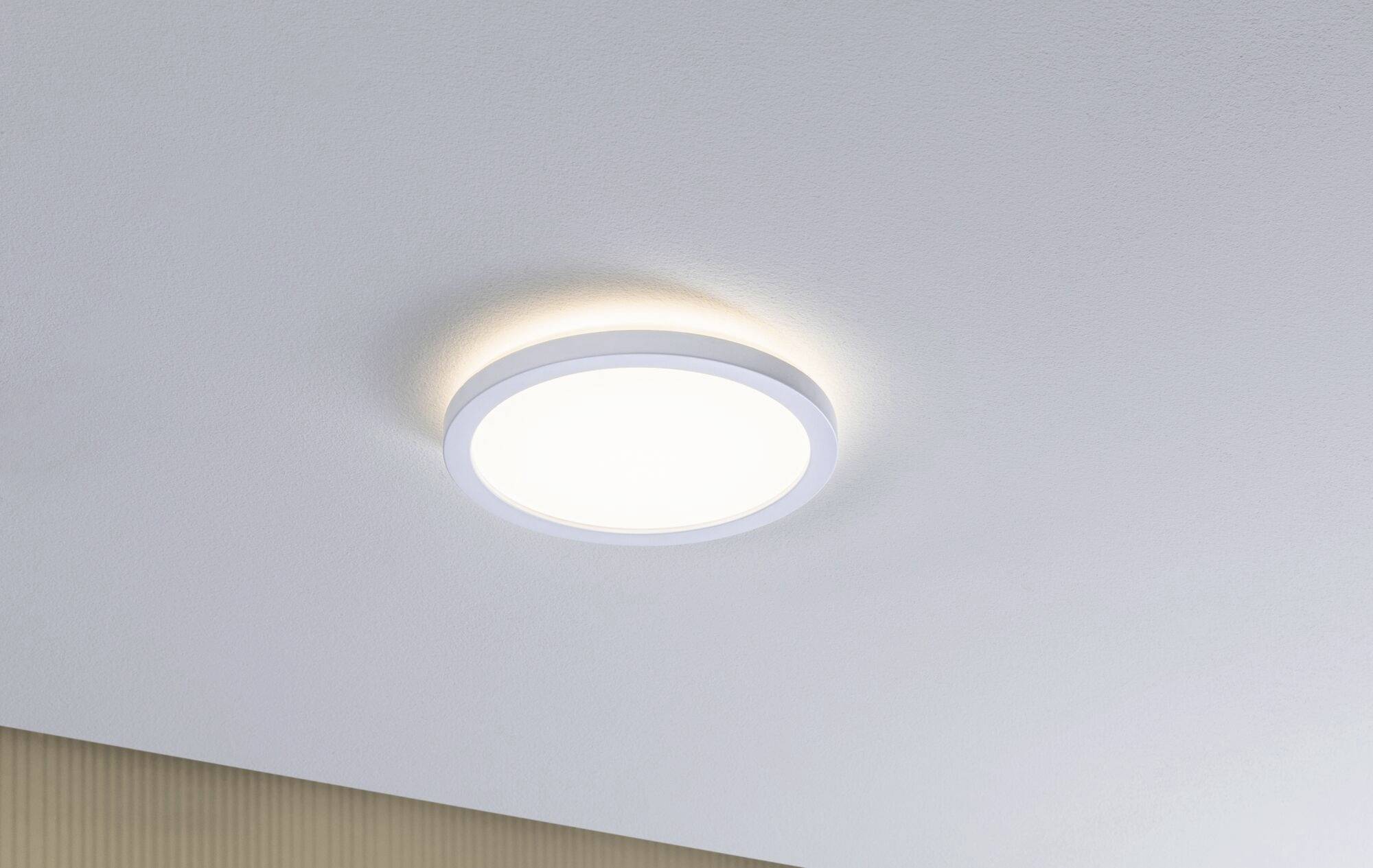 Luminaire plafonnier rond et blanc au design simple et moderne, monté sur un plafond lisse et gris clair. Il diffuse une lumière uniforme.