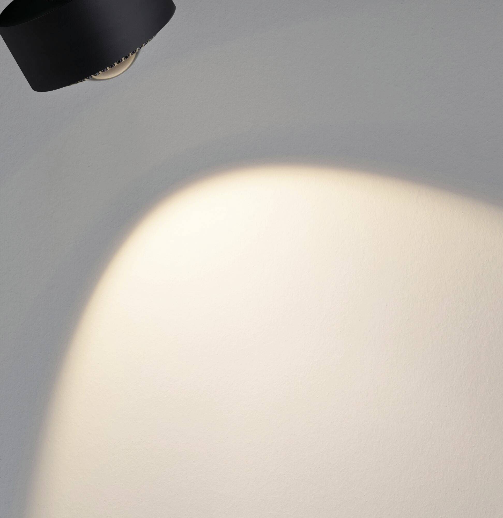 Une applique noire éclaire un point lumineux sur un mur blanc par ailleurs uniforme.