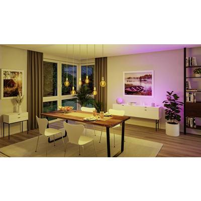 Une salle à manger avec une table en bois et des chaises blanches. Des lampes suspendues éclairent la pièce. De grandes fenêtres et des plantes en arrière-plan.