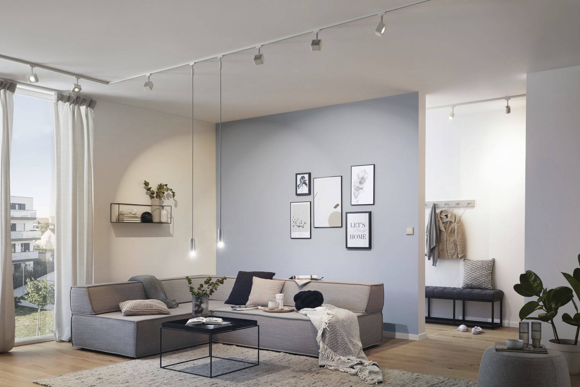 Un salon moderne avec un canapé d'angle, une table basse et un mur décoratif orné de cadres. La lumière du jour entre par la fenêtre.