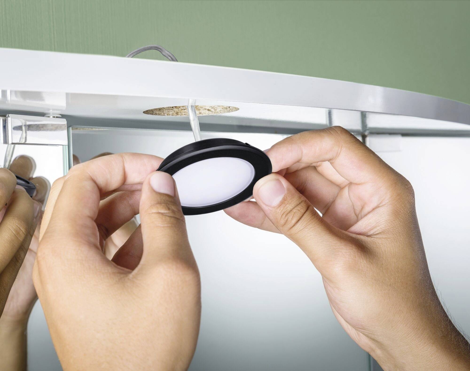 Des mains installent un luminaire LED circulaire dans un plafond. Des câbles et une ouverture ronde dans le miroir sont visibles.