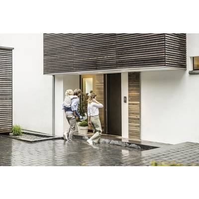 Une famille entre dans une maison moderne. Un homme porte un enfant, la femme ouvre la porte. Il pleut légèrement, et l'entrée est mouillée.